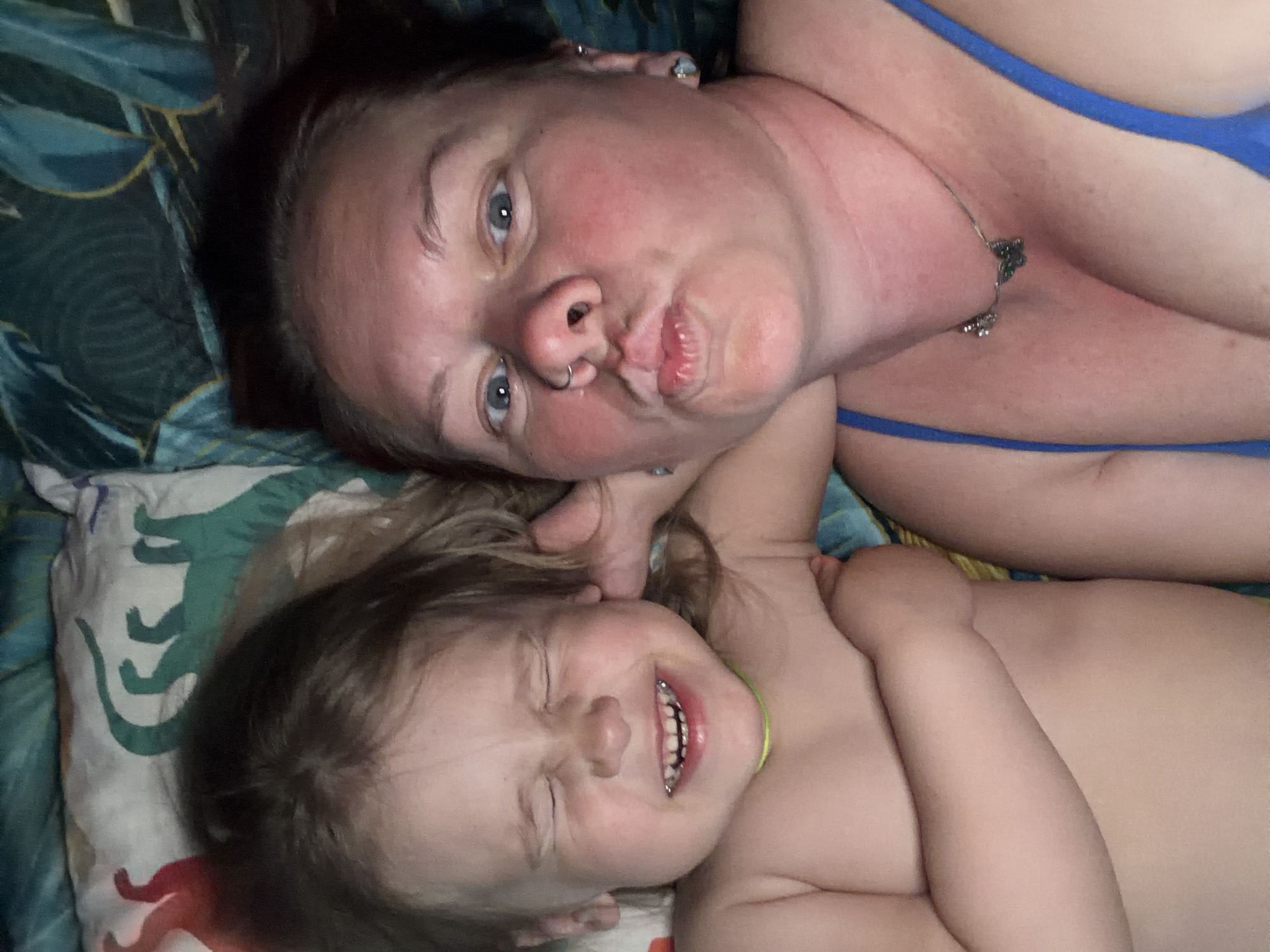 A selfie of a woman and a child lying down on a pillow and bedding. The woman is light-skinned with dark hair, blue eyes, and a nose ring. She is making a pouting facial expression. She is wearing a blue top and a necklace. The child is light-skinned with light brown hair and is smiling with eyes closed. The child is not wearing clothing. The pillow has a dinosaur pattern. The bedding has a blue and green leaf pattern. The location is not specified.