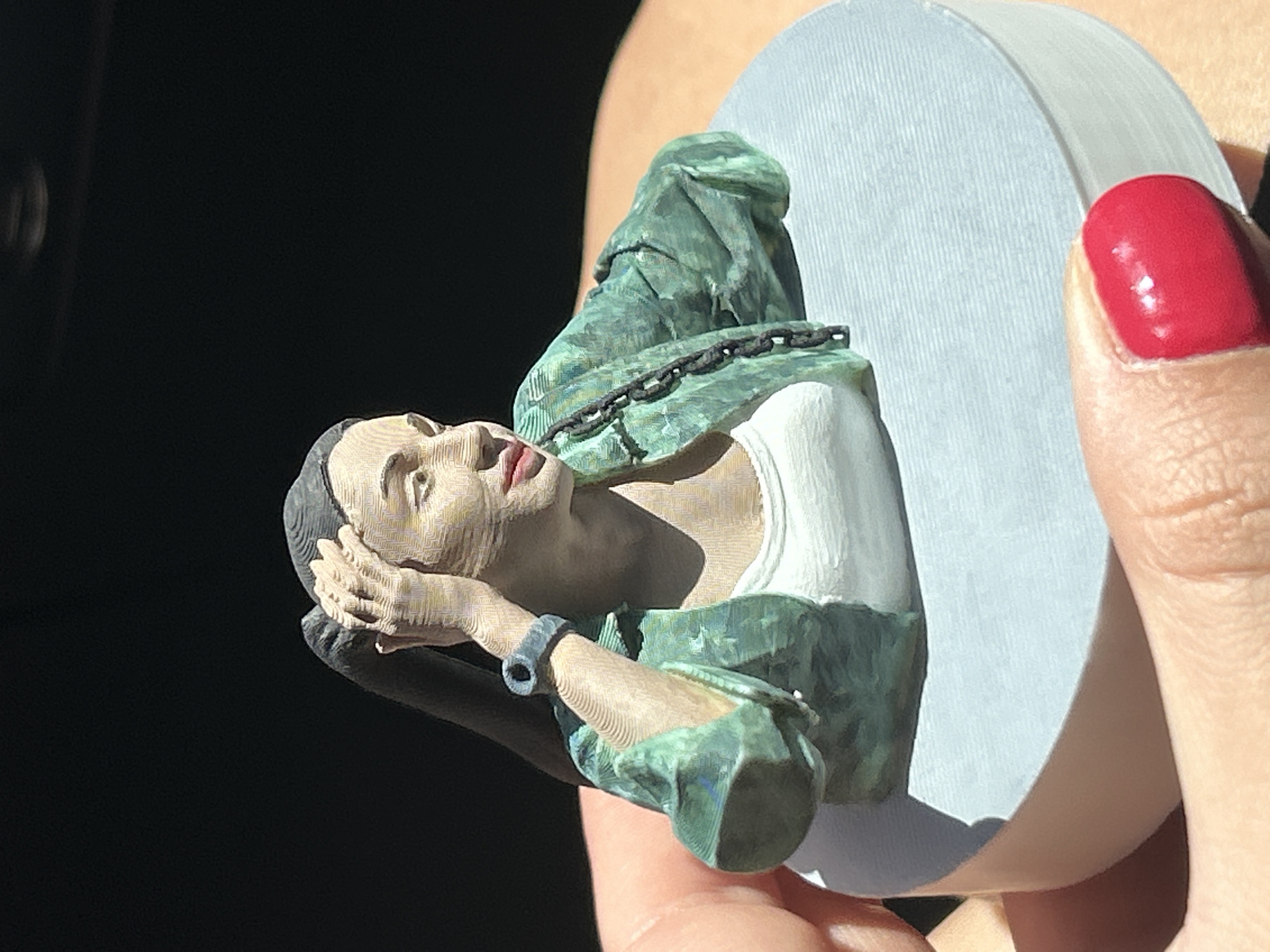 A close-up, full-body shot of a small, 3D-printed figurine of a woman is held between two hands. The figurine depicts a woman with dark hair pulled back, wearing a white tank top and an unbuttoned green jacket with a black chain detail. Her right hand is raised to her head, and she is looking to her right. The figurine is mounted on a light blue circular base. The background is dark and out of focus, with a bright light source from the upper left casting shadows. The hands holding the figurine have neatly manicured red fingernails and skin texture visible. The image is taken from a slightly elevated angle. The specific location in Lozen, Bulgaria, is not discernible from the image content.