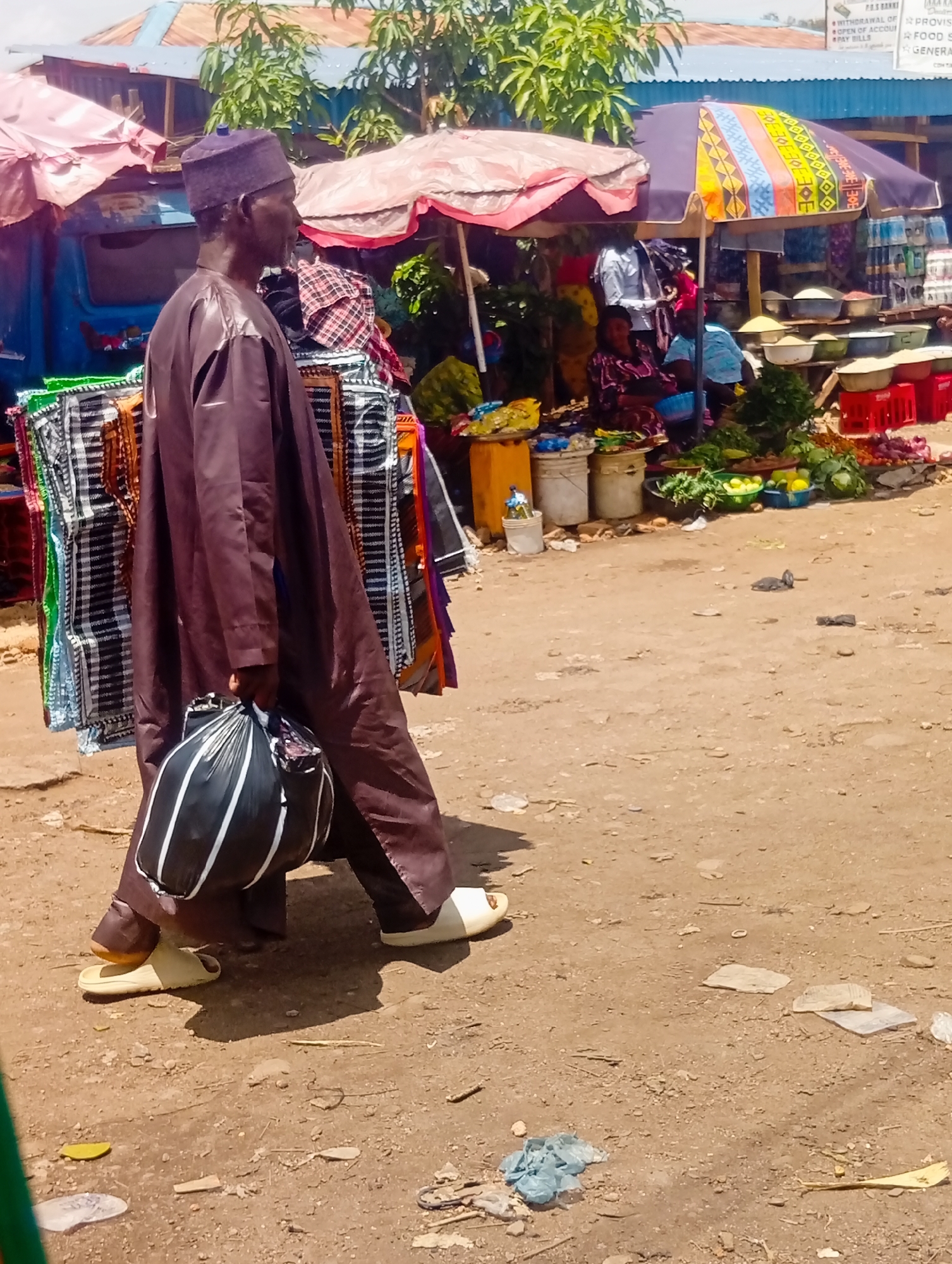 The image depicts a street scene in Bukuru, Nigeria. A man wearing a brown robe and a cap walks from left to right, carrying a black and white striped plastic bag in his left hand. He is wearing white slides on his feet. Behind him, a market stall is visible with various goods, including textiles, umbrellas, and produce. The ground is dusty and appears to be a dirt road. There are several other people in the background, partially obscured by the stall. The overall impression is that of a busy marketplace.