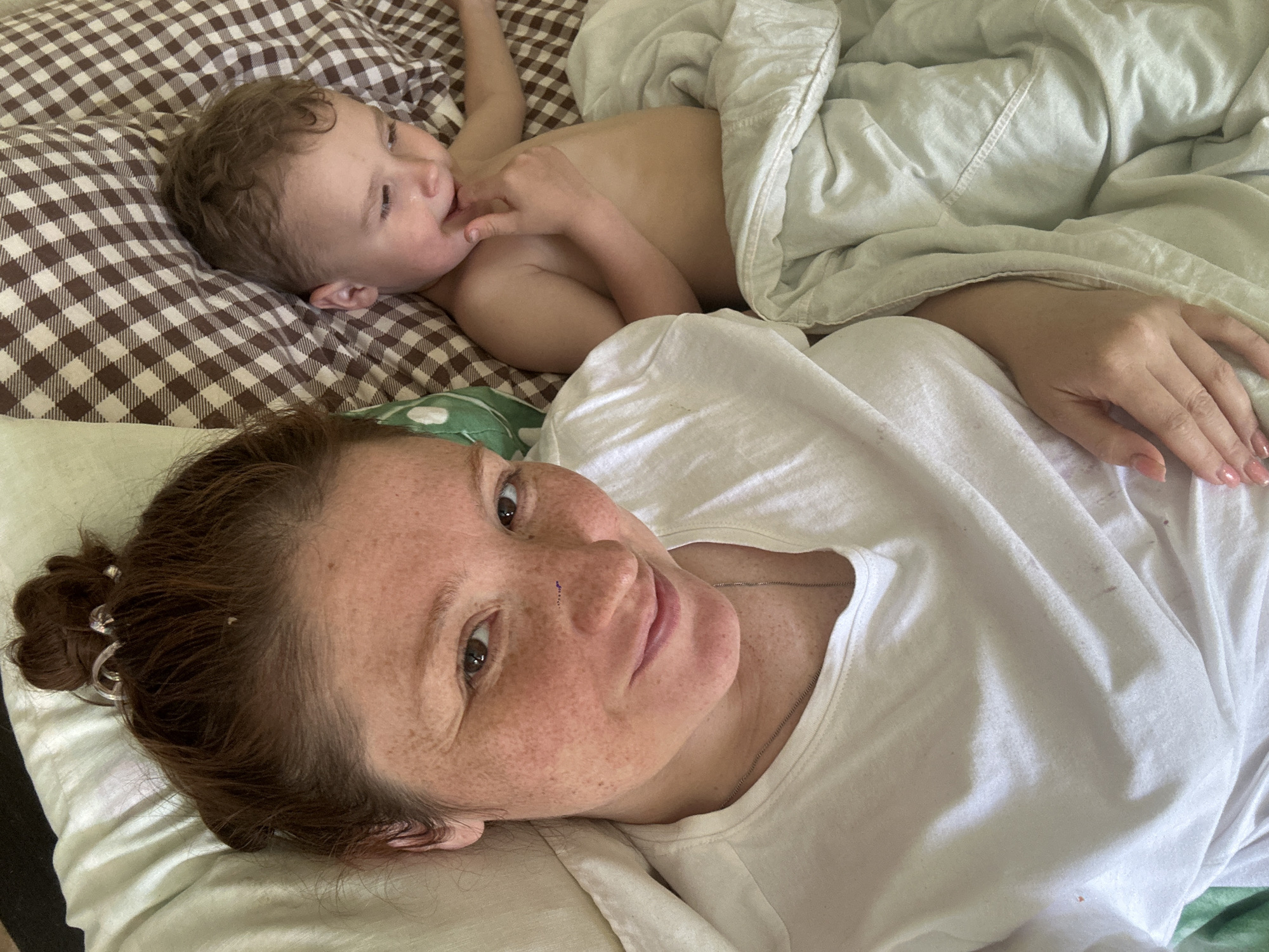 A fair-skinned woman with freckles and auburn hair pulled back in a messy bun is lying on a bed in what appears to be a domestic interior. She is wearing a white t-shirt and looking at the camera. Adjacent to her, a light-skinned toddler with curly hair is lying on their back on a brown and white checkered pillow, partially covered with a white blanket. The child has a hand near their mouth. The overall scene suggests a moment of rest or relaxation indoors. There is no evidence to indicate the location of Nizhny Novgorod, Russia.