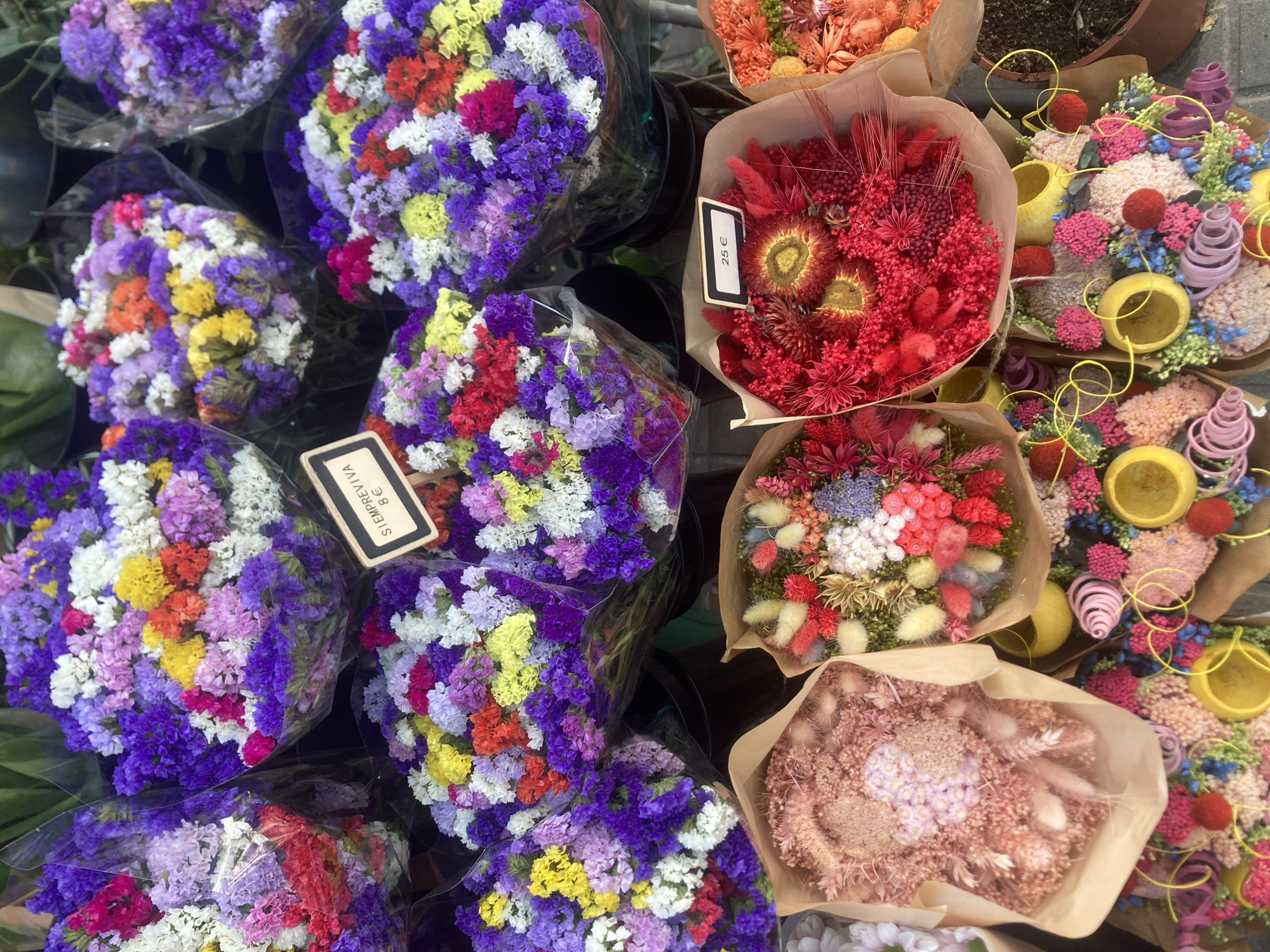 The image shows a colorful display of dried flowers arranged in bouquets for sale. The flowers are predominantly in shades of purple, pink, white, yellow, and red. Some bouquets are wrapped in clear plastic, while others are presented in brown paper. Two small price tags are visible: one reads "SIEMPREVIVA 8€" and another reads "25€". The scene appears to be an outdoor market stall, likely in Barcelona, Spain, given the presence of the Spanish language on the price tag. The background is blurred, but suggests a marketplace environment. No people are visible in the image.
