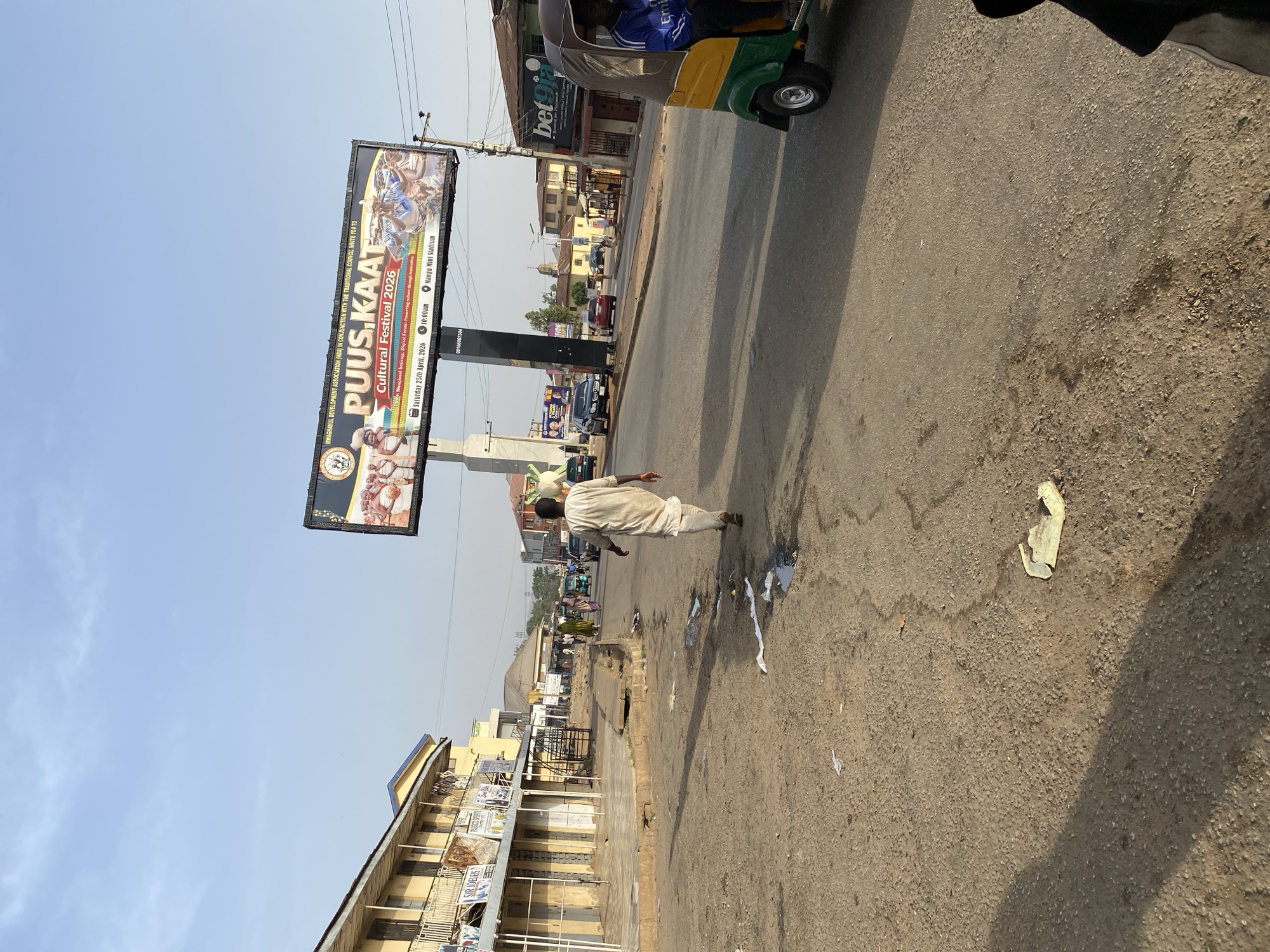 The image shows a street scene in Jos, Nigeria, featuring a large billboard advertising the "PUUS KAAT Cultural Festival 2026" at Nangu Mini Stadium on April 25, 2026. A person wearing light-colored traditional attire is walking along the side of the road. The road surface appears worn and dirty, with scattered debris. A three-wheeled vehicle, partially visible, is on the right side of the frame. Buildings line the street, including a structure with multiple advertisements. The sky is clear.