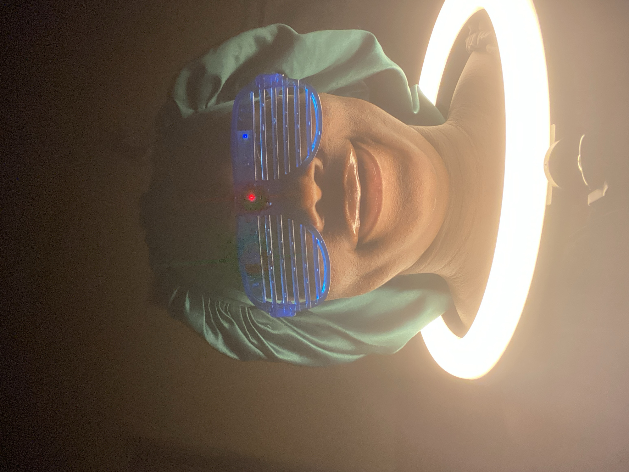 A person wearing blue shutter shades and a green head covering is centered in a dimly lit indoor space, with a ring light illuminating the right side of their face. The ring light and person are the primary subjects. The location is unknown, but presumed to be Maigizo, Nigeria.