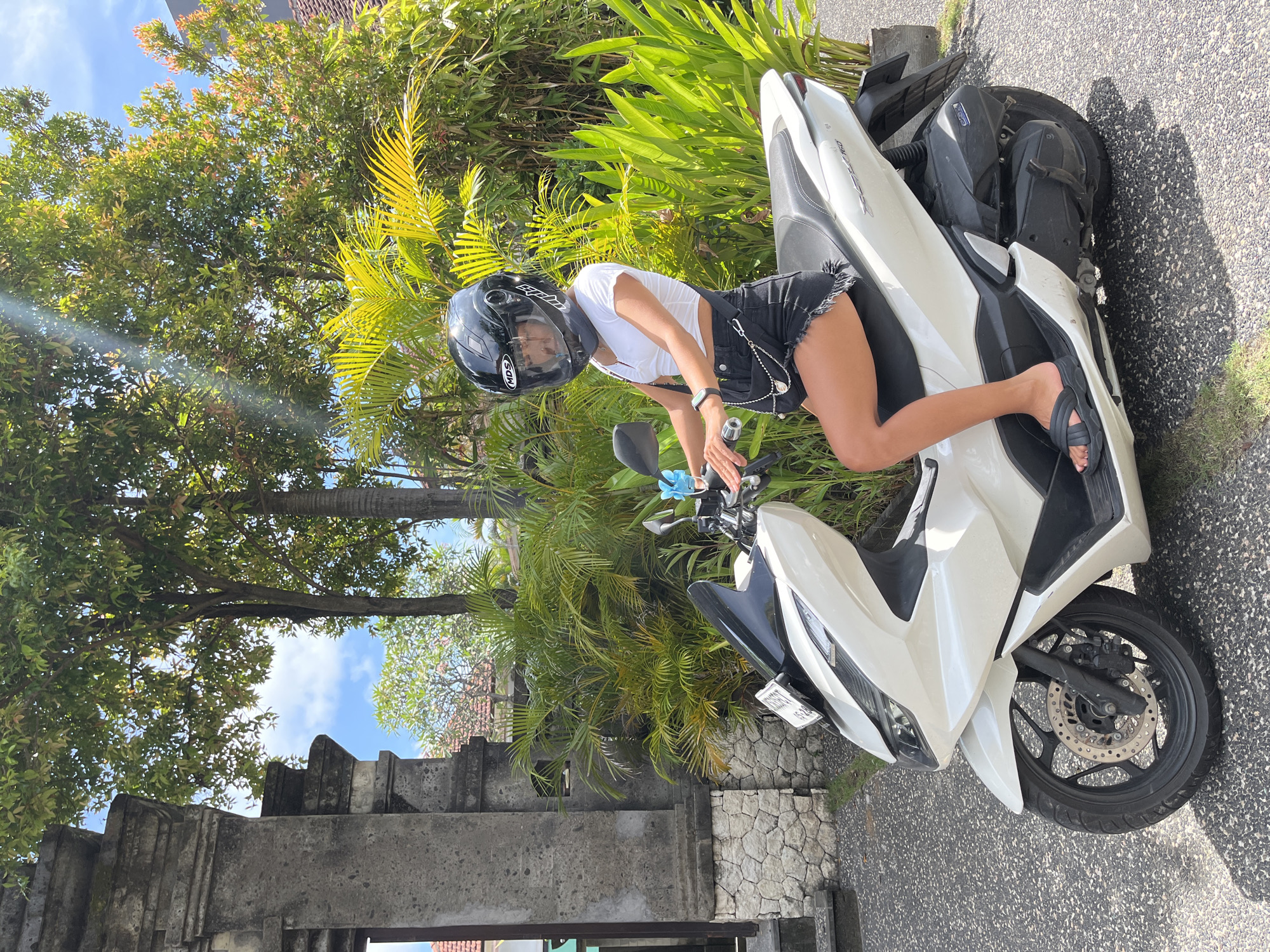 A young woman in a white shirt, black shorts, and a black helmet is leaning against a white and black scooter on a paved surface. She is wearing black sandals and a watch. The scooter has a license plate.
The scene is set outdoors amidst lush tropical vegetation, including palm trees. In the background, there is a stone structure. The location appears to be Kuta Selatan, Indonesia.