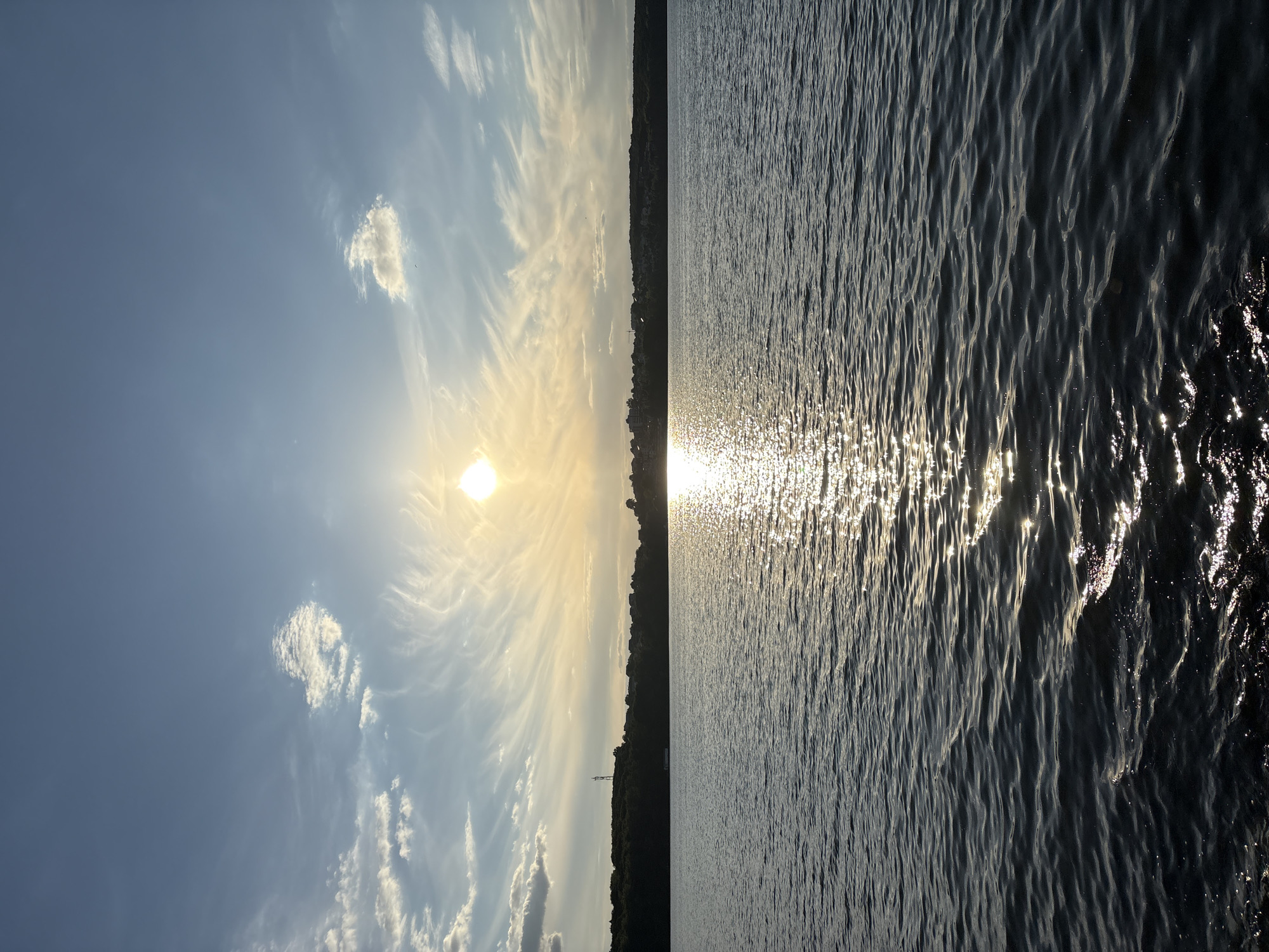 The image depicts a wide view of a body of water, likely a lake or a large river, under a bright sky. The sun is visible near the center of the frame, casting a strong reflection on the water's surface, creating a shimmering path of light. The water is slightly rippled, indicating gentle movement. In the background, a silhouetted treeline with some scattered buildings and a tall antenna or tower can be seen along the horizon. The sky is a clear blue with some wispy white clouds, particularly around the sun, suggesting it might be late afternoon or early evening. The overall impression is one of a serene, natural landscape. There are no people or animals visible in the image. The location is identified as Ternopil, Ukraine.