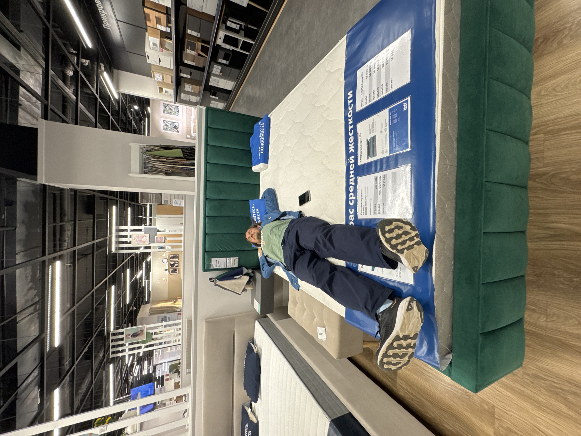 The image depicts a person lying on a mattress in a store, likely a furniture or bedding store. The person is lying face-up, wearing jeans, a green t-shirt, and a denim jacket. Their feet are visible, shod in running shoes. A mobile phone rests on the mattress near them.
The mattress is displayed with a green headboard and is covered in white fabric. A blue tag is attached to the mattress with text in Russian: "бас средней жесткости" and "ПОЖАЛУЙСТА". Additional product information and pricing are visible on a blue label affixed to the mattress, including text in Russian, such as "Mildex Energy Life" and "Hoff".
The store environment is visible in the background, showing aisles with shelves filled with various products. The lighting is bright and overhead. A black metal structure is visible near the ceiling.
The location is likely in Izhevsk, Russia, based on the text in the image and the Hoff brand.
