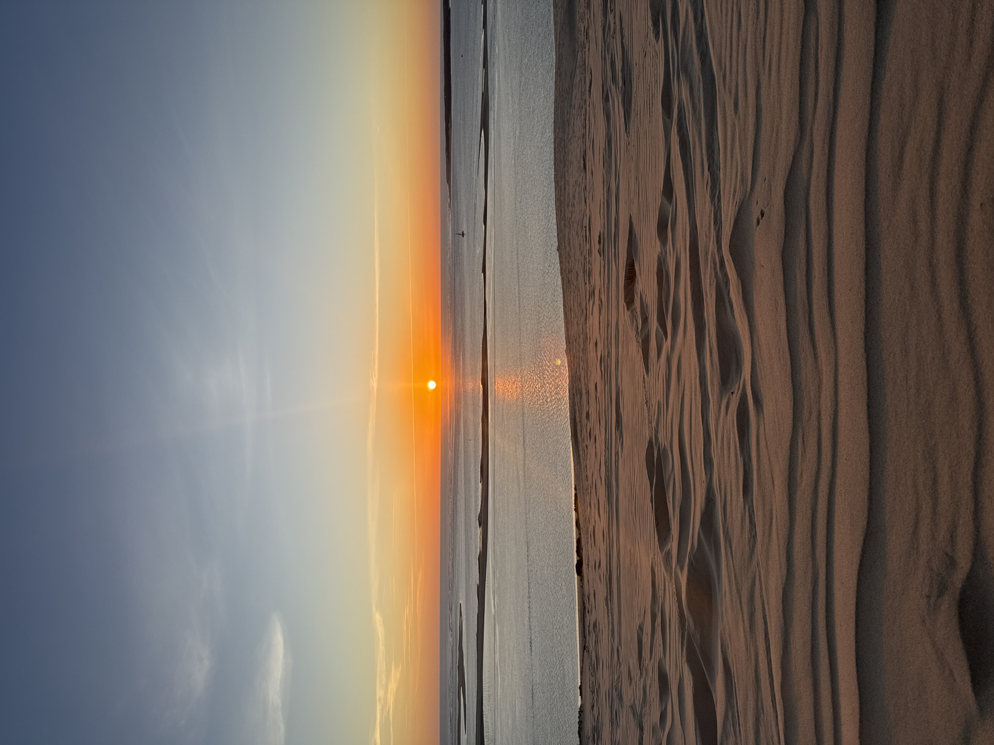 This image depicts a serene sunset over the Dune of Pilat, located in La Teste-de-Buch, France. The foreground is dominated by the undulating, rippled sand dunes, casting long shadows that emphasize their textured surface. The middle ground features a body of calm water, likely the Arcachon Bay, reflecting the warm hues of the setting sun. A narrow strip of land or sandbar is visible in the bay, with some vegetation or structures. In the distance, the horizon is marked by a faint line of land. The sky transitions from a pale blue at the top to vibrant shades of orange and yellow near the horizon, where the sun, a bright orange orb, is setting. A few wispy clouds are scattered across the upper sky, and a small sailboat can be seen on the water. The overall scene is tranquil and picturesque, showcasing the natural beauty of this popular tourist destination.