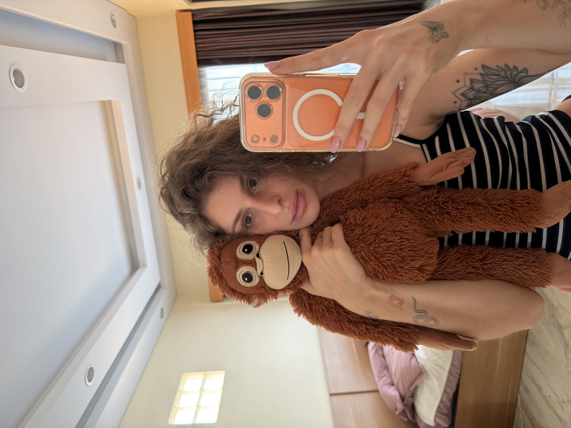 A young woman is taking a selfie in a bedroom. She is holding a stuffed monkey in her left arm and her orange-cased phone in her right hand. She is wearing a black and white striped tank top. The room has a bed with pink and white bedding, and a window with frosted glass. There are tattoos on her arms, including a snake and a butterfly. The ceiling has recessed lighting and a dropped ceiling design.