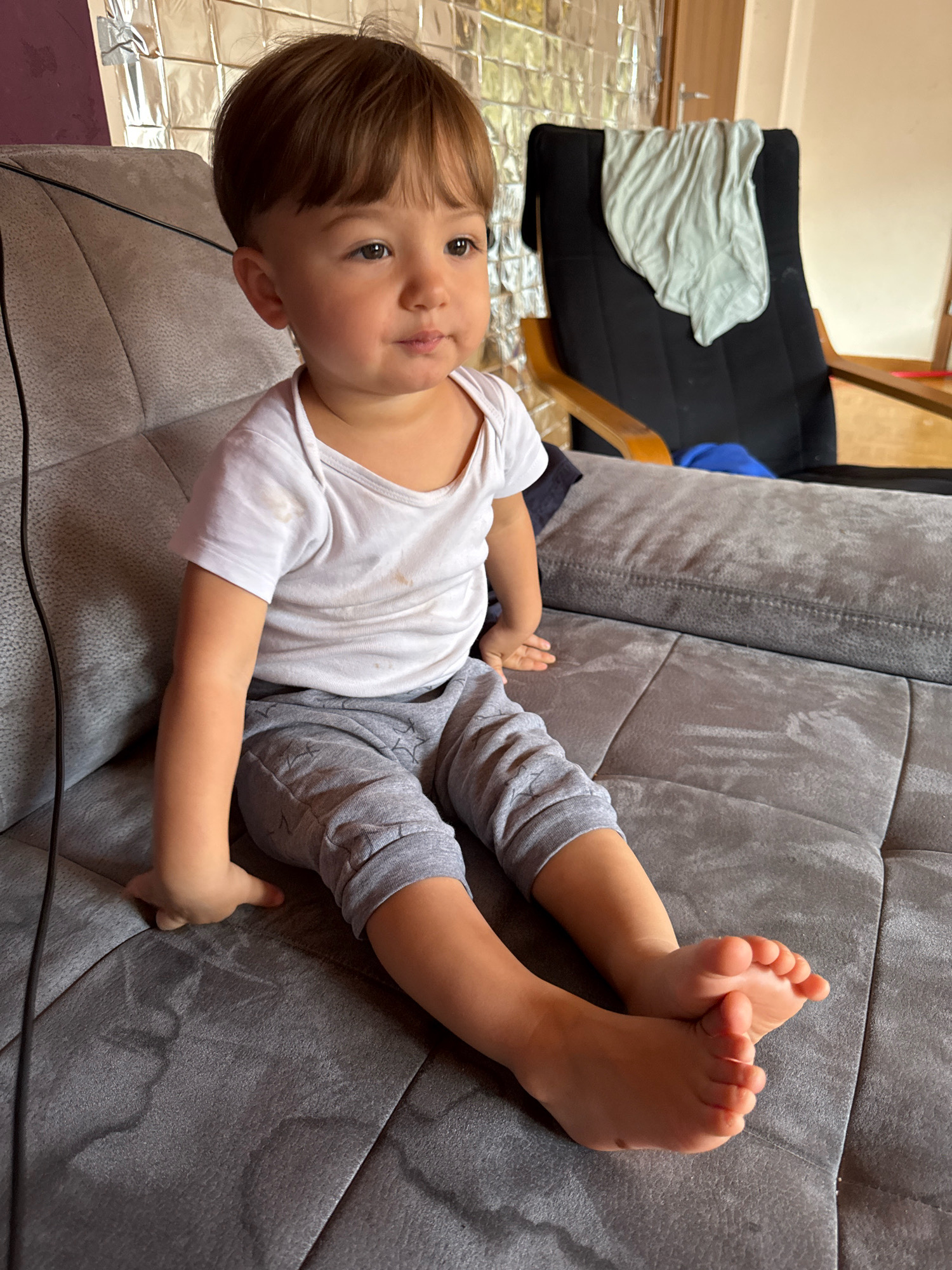 A young child with brown hair and light skin is sitting on a gray sofa. The child is wearing a white t-shirt and gray pants. The child's feet are visible and are resting on the sofa. The background shows a wall with a metallic-looking texture, a black chair with a light blue cloth draped over it, and a wooden door. The overall scene appears to be indoors, likely in a residential setting within Überherrn, Germany.