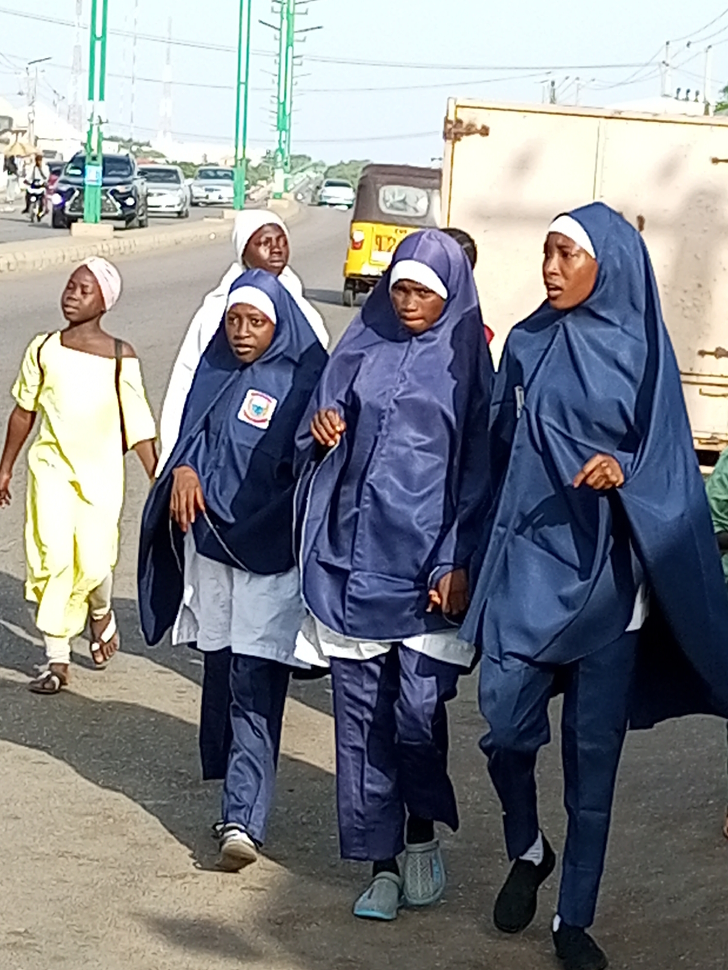 In Gombe, Nigeria, a group of young people, likely students in uniform, are walking on a paved road. The individuals are wearing traditional Islamic attire, including hijabs and long, loose-fitting garments in shades of blue and white. One person in the foreground wears a vibrant yellow outfit. They appear to be walking together as a group, with some holding hands. The background shows a busy street with various vehicles, including cars, a motorcycle, and a yellow tricycle, suggesting an urban environment. Traffic lights and utility poles are visible, indicating infrastructure. The time of day appears to be daytime, with shadows cast on the road. The overall scene depicts a daily commute or movement within the city.