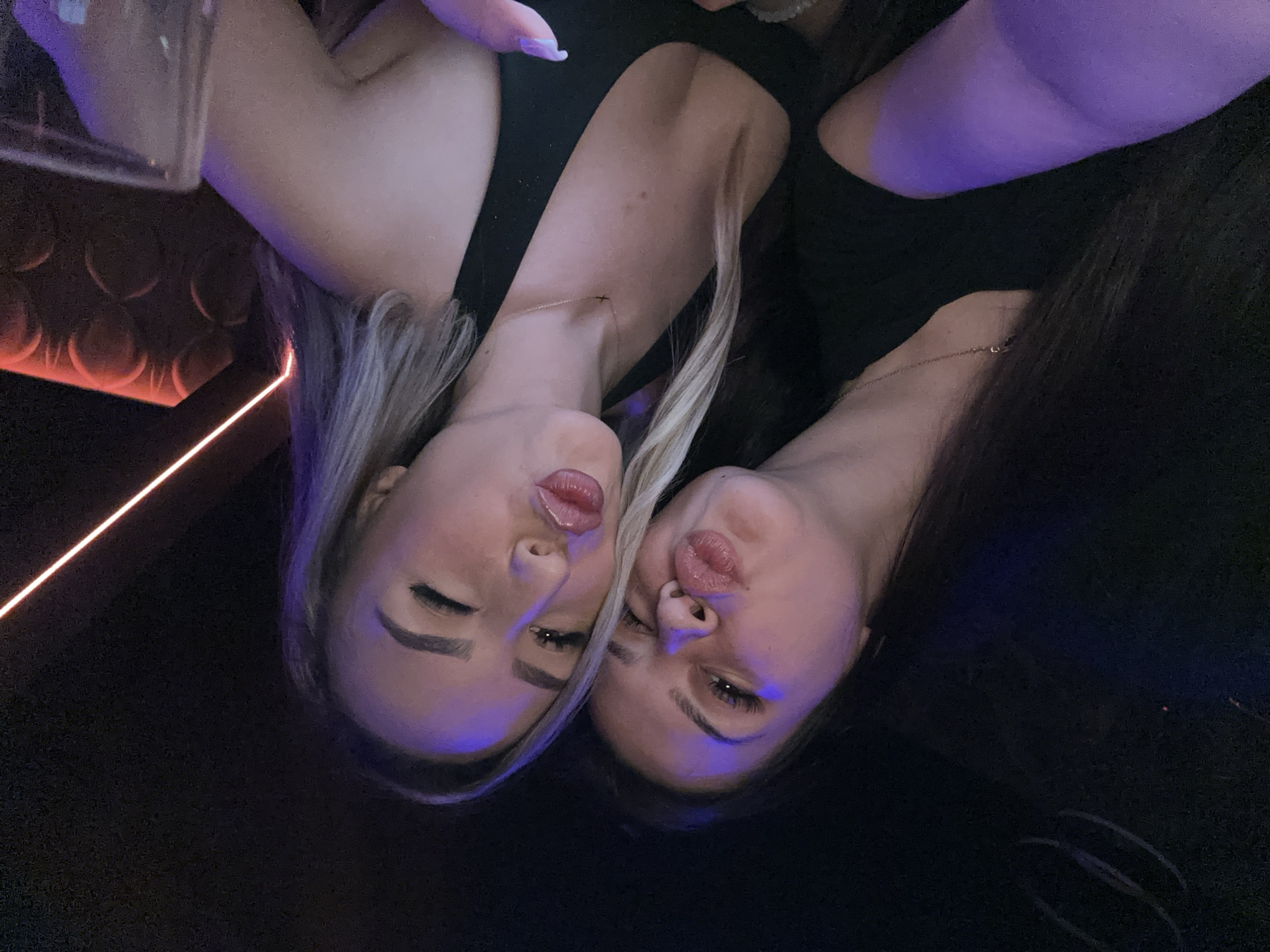 Two women are close together, posing with pursed lips. The woman on the left has blonde hair, and the woman on the right has dark brown hair. Both are wearing black tops and necklaces. The background is dark, with a faint red light source and a patterned wall visible. The image appears to have been taken indoors, possibly in a bar or nightclub. The location is not discernible as being in Sliven, Bulgaria.