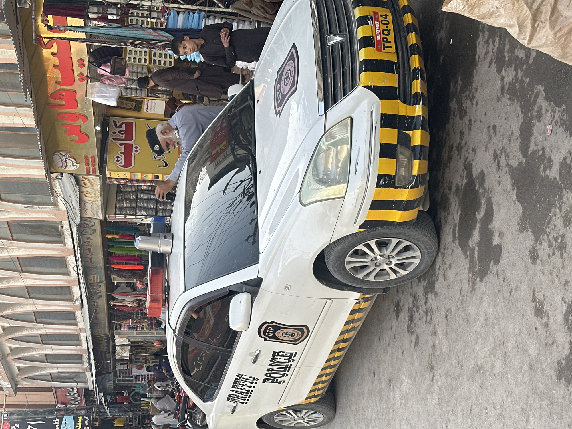 A white police car, marked "TRAFFIC POLICE" and featuring the "QTP" (Quetta Traffic Police) logo, is prominently parked on a bustling street. The car has distinct black and yellow diagonal stripes on its front bumper and lower body, along with red and blue siren lights on its roof. The license plate holder reads "TRAFFICE POLICE TPQ-04".
The street is a lively market area, with numerous shops lining the left side. Several people are visible, including a man with a white beard and a dark cap, dressed in a light blue shirt, who is leaning towards the side of the police car. Other individuals, including a young boy in a dark outfit, are seen browsing or walking near the shops in the background.
Shop signs are written in Arabic/Urdu script, with visible text including "د خان اوران", "جائے نماز" (prayer mat), "عبد الله", "كيپ هاوس" (Cap House), "كفایت" (Kifayat), and "الخدمت". The buildings have arched architectural details. The scene takes place during the day, under what appears to be an overcast sky, as indicated by the soft lighting and lack of harsh shadows. The road surface is concrete or asphalt, showing some moisture or stains. The overall setting suggests a vibrant urban market in Quetta, Pakistan.