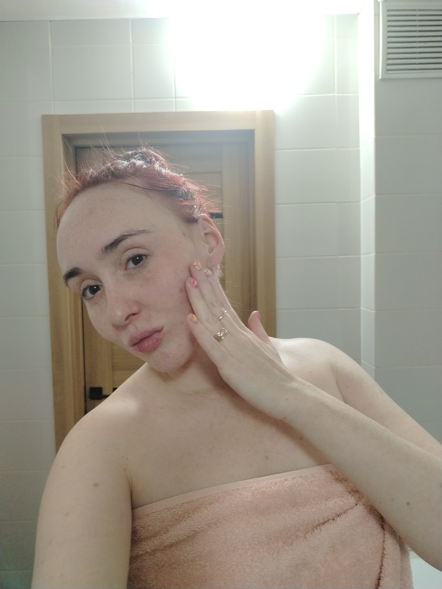 The image displays a self-portrait of an adult individual, appearing female, with fair skin and reddish hair pulled back. The subject is wrapped in a peach-colored terry cloth towel, revealing the bare shoulders and upper chest. Visible features include numerous freckles on the face, shoulders, and arms, alongside scattered reddish blemishes on the cheeks and upper chest. The subject's left hand is raised, with fingers lightly touching their left cheek. This hand features multicolored patterned fingernail polish and a gold-colored ring with a central design on the ring finger. The individual's lips are slightly puckered, and their gaze is directed towards the camera.
The scene is an interior space, characterized by white rectangular ceramic tiled walls. In the background, to the left of the subject, a section of a light-colored wooden door frame with a dark handle is visible. To the upper right, a white rectangular ventilation grate is installed on the wall. The illumination is bright, causing a slight overexposure in the upper right corner of the frame. The precise location within Izhevsk, Russia, is not identifiable from the visual content.