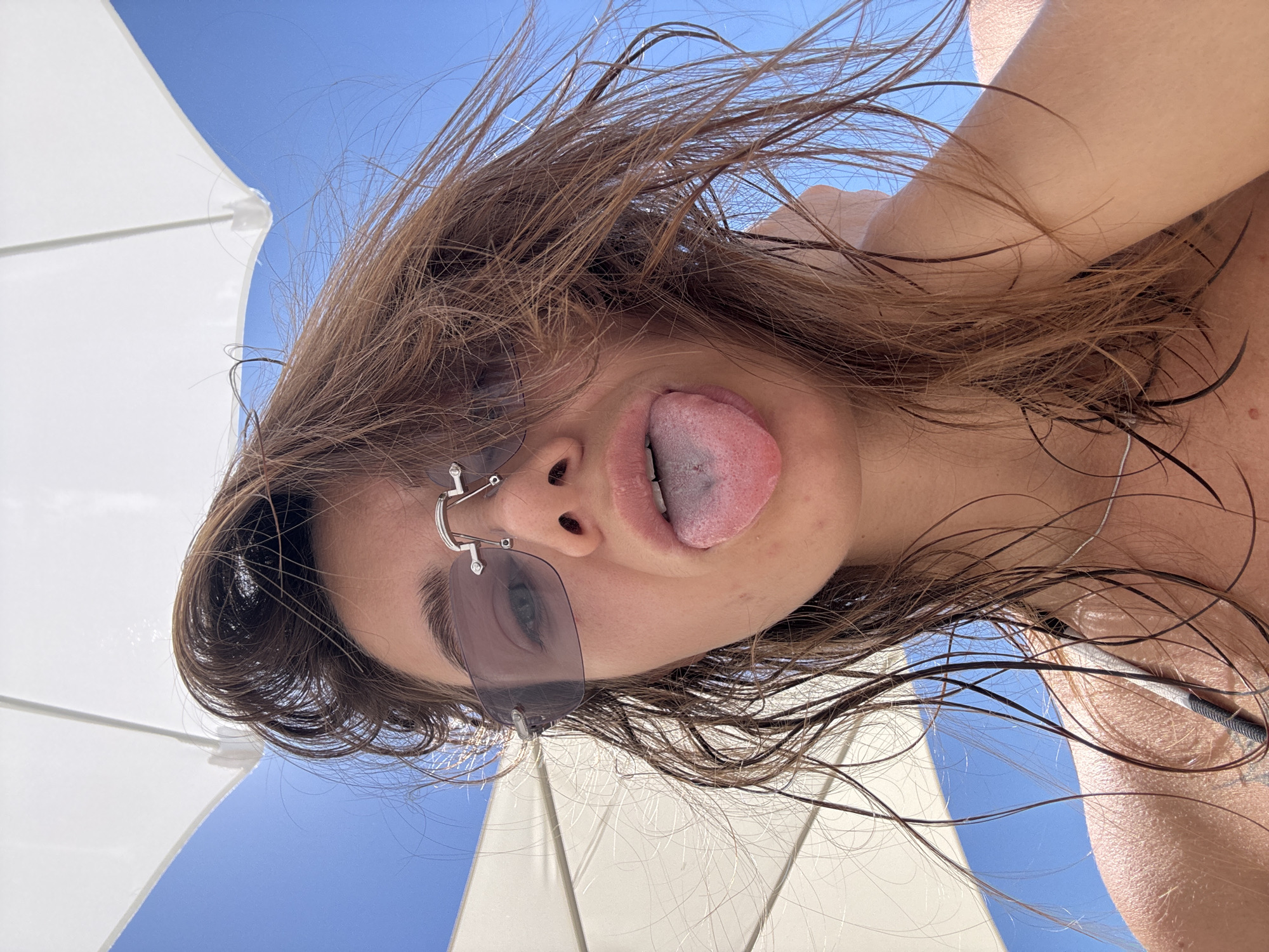 An upward-facing photographic image depicts a female individual centered beneath a white parasol against a clear blue sky in Boreti, Montenegro. The individual has wet, brown, windswept hair and is wearing rimless rectangular sunglasses with dark lenses. Her mouth is open, and her tongue is extended, displaying a small silver piercing. The tip of her tongue exhibits a slight blue-purple discoloration. Her skin appears tanned and glistens with moisture. A thin silver necklace is visible on her neck, and the dark strap of a garment, possibly a swimsuit, is visible on her left shoulder. Her left hand is raised, with fingers positioned behind her head. The white parasol occupies the upper portion of the frame, partially obscuring the sky, with its internal structure visible.