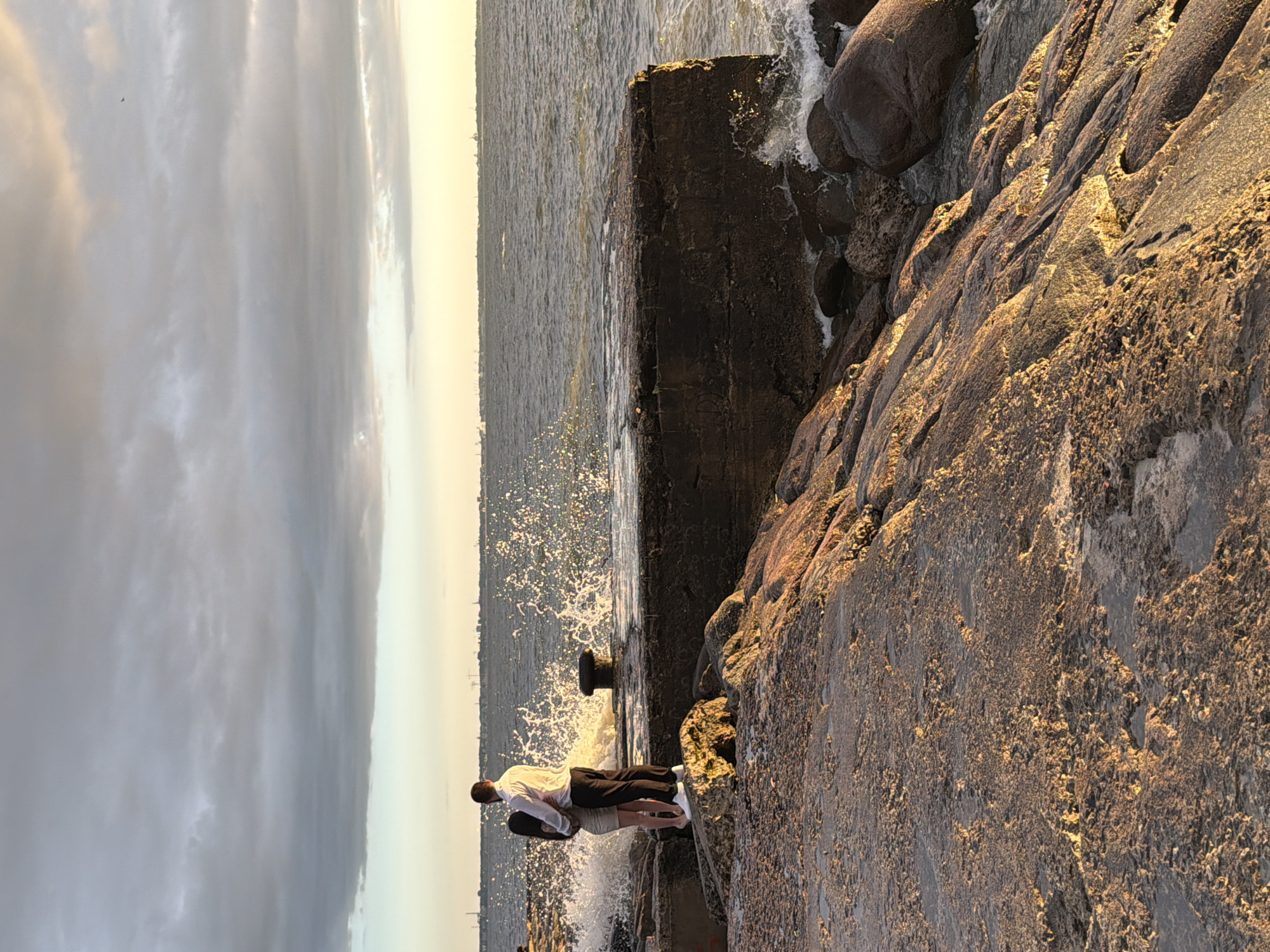 This is a medium-angle, full-shot photograph of a couple embracing on a rocky shoreline with a concrete pier in the background, at sunset in Tallinn, Estonia. The couple, a man in a white dress shirt and black pants and a woman in a short, light-colored dress, stand facing away from the camera, looking out at the sea. Waves crash against the pier, creating a spray of water. The sky is filled with dramatic, cloudy formations tinged with the warm hues of the setting sun. The foreground is dominated by textured, sunlit rocks, with the sea and a distant, hazy city skyline in the background. The image captures a moment of romantic solitude against a backdrop of natural beauty and urban presence.