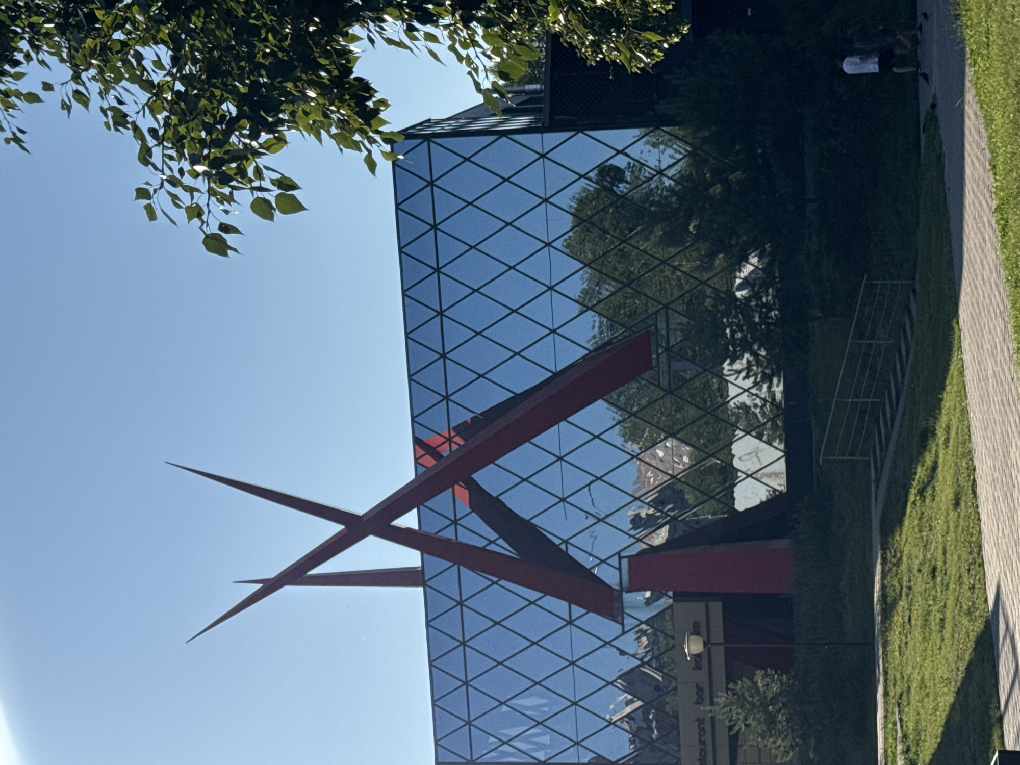 A contemporary building with a geometric glass facade stands under a clear blue sky. Two large, red, angular sculptures pierce the sky from atop the structure. The glass reflects trees and buildings, creating a distorted, diamond-patterned surface. In the foreground, a grassy area leads to a paved walkway. Two individuals, a man in a white t-shirt and black shorts and another person in a dark shirt, walk on the walkway. A lamppost stands near the building's entrance, and some signage is visible, including "staurant" and "bar." This scene is located in Nizhny Novgorod, Russia, likely at the building known as the "Pyramid" or Nizhny Novgorod Kremlin's exhibition hall.