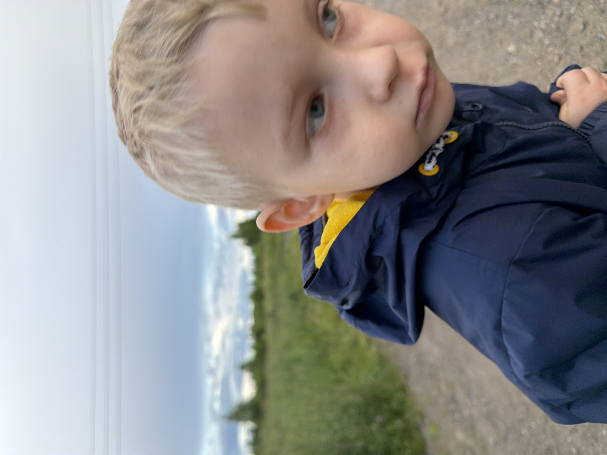 The media file is a close-up, rotated photo of a young child with blonde hair. The child is wearing a dark blue jacket with yellow accents on the zipper pulls and collar. The child's face is turned towards the camera, with a slight tilt to their head. They have blue eyes and fair skin. The background is blurred, showing a grassy field and a cloudy sky, suggesting an outdoor setting, possibly in the countryside. The lighting suggests it might be daytime, possibly late afternoon or early evening, given the soft light. The child's expression is neutral or slightly curious. There is no visible text in the image. The provided location context is "Чёрная, Russia".