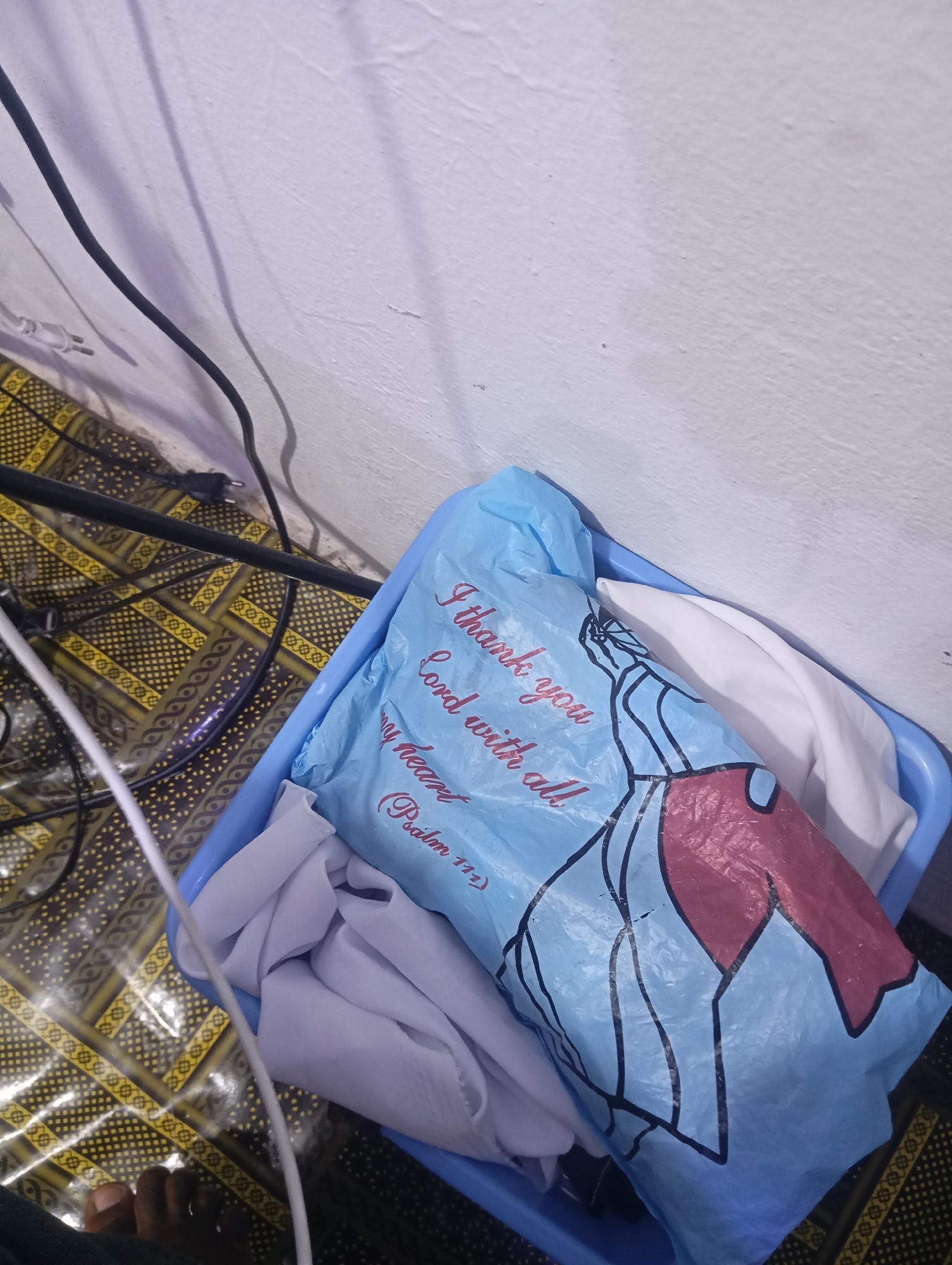 A blue plastic bag with red and black text and graphics is placed inside a blue plastic basin. The text on the bag reads "I thank you Lord with all my heart (Psalm 112)." The bag appears to contain white folded fabric. The basin is situated on a floor covered with a patterned mat that has yellow and black geometric designs. Black and white electrical cords are visible on the mat and near a white wall. A person's foot, with dark skin and visible toes, is in the lower left corner of the image. This scene is located in the city of Jauro Yinu, Nigeria.