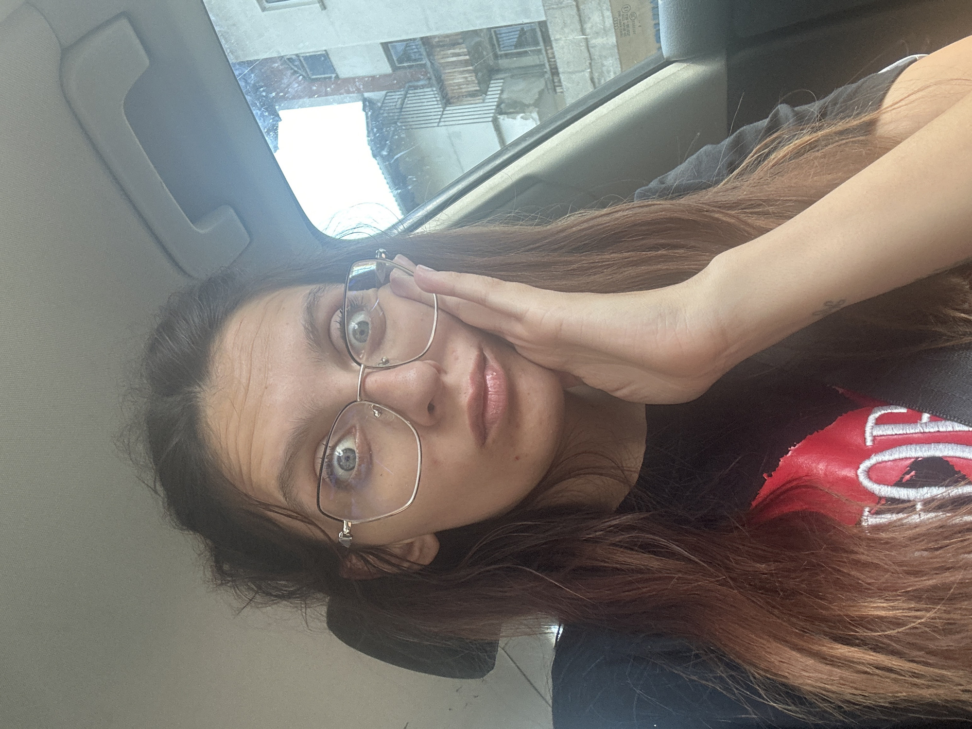 The image shows a young woman with long, brown hair wearing glasses and a black t-shirt with a red graphic. She is sitting in the passenger seat of a car, with her hand pressed against her cheek, and her eyes are wide open. Through the car window, a street scene in Tryavna, Bulgaria is visible, featuring old buildings with balconies and windows. The sky is overcast.