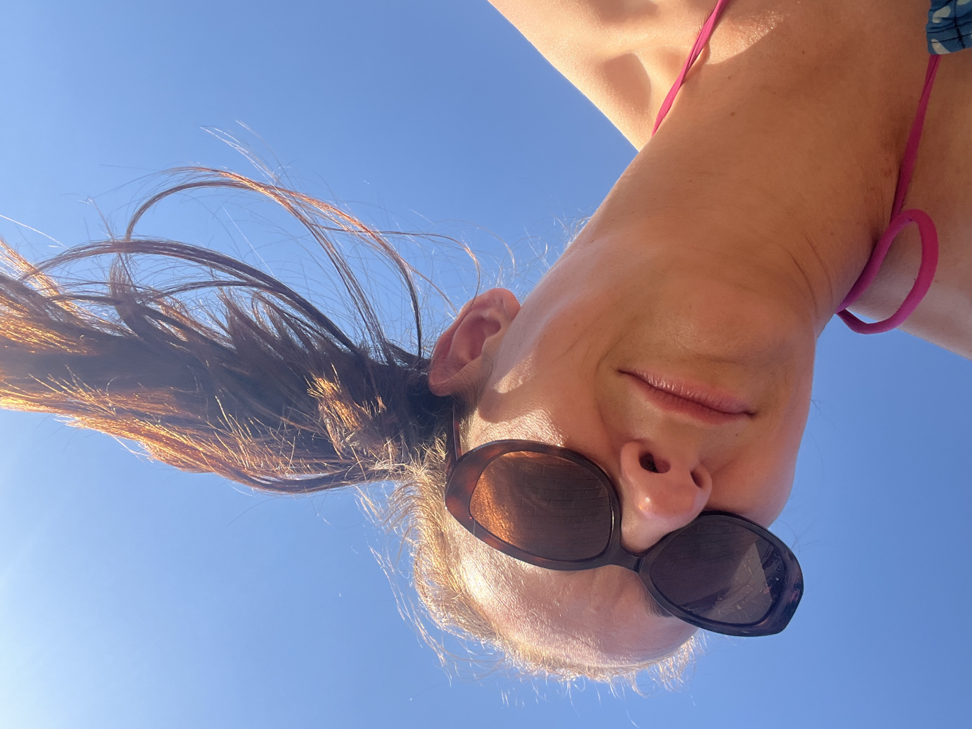 This is a close-up, upside-down selfie of a woman against a bright blue sky. She is wearing dark tortoiseshell sunglasses and a pink bikini strap is visible around her neck. Her hair, which appears to be dark brown with reddish highlights, is long and flowing, catching the sunlight. The lighting suggests it is daytime, likely sunny. The perspective of the shot, looking up at the woman from below against the sky, suggests she might be lying down or hanging upside down, perhaps at a beach or pool. The overall mood is casual and carefree, implied by the summery attire and the outdoor setting. There is no visible text in the image.
