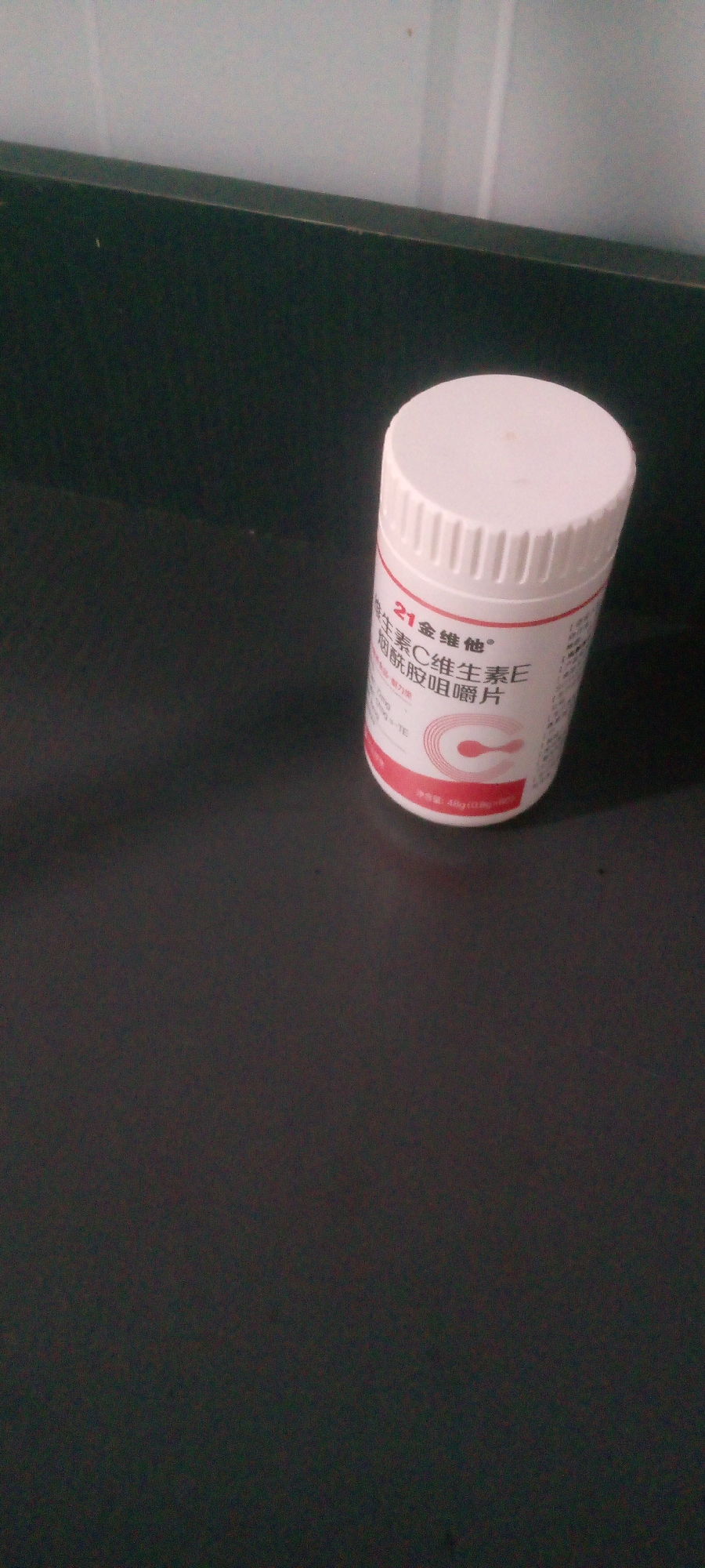 The image shows a white bottle of vitamin supplements with a red and white label. The label has Chinese text and a prominent "C" logo. The bottle is standing on a dark surface, possibly a table, with a blurred dark green surface in the background. There is no indication of the location being in Indo, Nigeria; the environment appears to be indoors. The image is a close-up of the bottle, with the rest of the surroundings out of focus.
