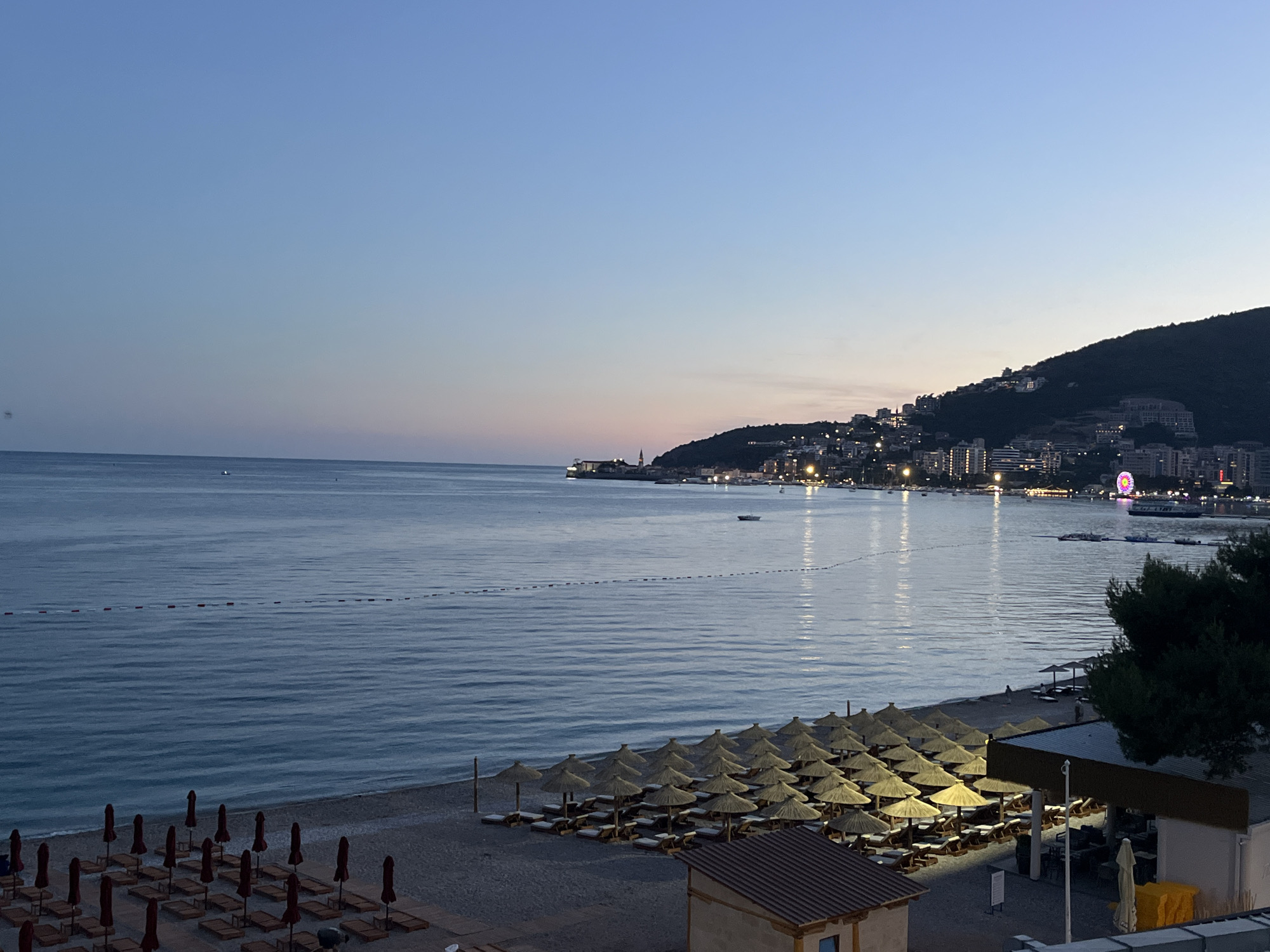 The image presents a panoramic view of Budva, Montenegro, at twilight, observed from an elevated position overlooking a bay. The sky transitions from a light blue overhead to a pale pink and orange gradient at the horizon where the sun has recently set. The calm, dark blue-grey sea exhibits gentle ripples and reflects the fading light and artificial illuminations from the distant city. A line of red-and-white buoys extends horizontally across the mid-ground of the water. Several small, indistinct boats are visible on the water's surface in the distance.
In the foreground, a wide beach occupies the lower portion of the frame. On the left side of the beach, numerous dark red, closed beach umbrellas are arranged in rows alongside corresponding sun loungers. On the right side, an array of open, straw-thatched beach umbrellas provides shade for sun loungers, illuminated by artificial lighting from unseen sources. Two low-rise structures with dark tiled roofs are present on the beach; one on the far left is partially obscured, while the one on the right appears to be a lit beach bar or restaurant. A cluster of dark green trees frames the right foreground.
Across the bay, the city of Budva is visible, its lights illuminating the coastline and the adjacent mountain. To the left, the distinct outline of Budva's Old Town peninsula is apparent, topped by a prominent red-roofed church tower (likely Sveti Ivan Church). The city's buildings stretch along the waterfront and ascend the slopes of a dark, tree-covered mountain that dominates the background on the right. A large, brightly lit Ferris wheel, displaying vibrant pink, purple, and yellow lights, stands out distinctly near the water's edge on the right side of the cityscape. Streetlights and building illuminations are scattered across the urban landscape extending up the mountain. One or two indistinct figures are visible on the beach near the open umbrellas on the right.