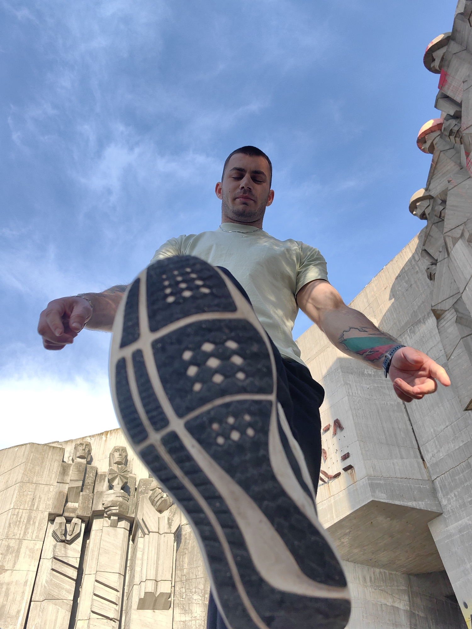 A low-angle shot captures a young man in an active pose against the backdrop of a monumental concrete structure and a clear blue sky. The central subject is a man with short dark hair, wearing a light-colored t-shirt and dark bottoms. He has a muscular build, and a prominent tattoo on his left forearm displays the colors of the Bulgarian flag (white, green, red) within a geographic outline. His eyes are closed, conveying a serene or contemplative expression.
The most striking element in the foreground is the sole of a dark athletic shoe, belonging to the man's raised left leg, which points directly towards the camera. Its intricate tread patterns are clearly visible.
Behind the man, a colossal, brutalist concrete monument dominates the background. This structure features angular, stylized figures carved into its facade, appearing to depict soldiers and possibly other human forms, characteristic of Soviet-era memorial art. The monument rises majestically against the sky, suggesting its immense scale and historical significance. Faint reddish marks, possibly graffiti, are visible on some parts of its concrete surface.
The setting is outdoors during the daytime under a bright blue sky with scattered white clouds, indicating fair weather. The low-angle perspective emphasizes the height of both the man's pose and the imposing monument. No discernible text is clearly visible in the image. The architectural style strongly suggests a location in Varna, Bulgaria, consistent with monuments like the Monument of the Bulgarian-Soviet Friendship.