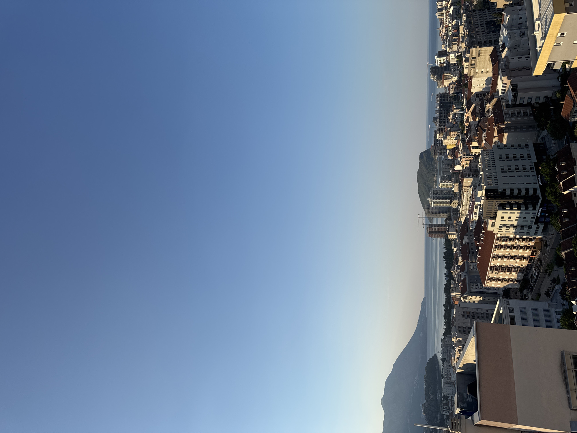 This is an aerial, high-angle, full shot of Budva, Montenegro, taken on a clear, sunny day with a bright blue sky. The city, nestled beside the Adriatic Sea, is a sprawling urban landscape characterized by numerous buildings of varying heights and architectural styles, predominantly residential. The foreground is dominated by the rooftops of apartment buildings, some with reddish-brown tile roofs and others with flat, modern designs.
In the middle ground, the cityscape continues with a dense collection of buildings, many appearing to be under construction, indicated by cranes and scaffolding. The buildings are a mix of white, grey, and beige, with some featuring balconies and darker-toned windows. The urban fabric transitions towards the coast, where the blue of the sea is visible.
The background features the distinctive geographical landscape of Budva. To the left, a large, verdant mountain range slopes down towards the sea. In the distance, a small, rocky island, possibly Sveti Nikola Island, is visible, covered in greenery and situated offshore. The calm sea stretches towards the horizon, meeting the clear sky. The overall impression is a vibrant coastal city with a strong presence of both natural beauty and ongoing development. No people or specific actions are discernible in this wide cityscape view.