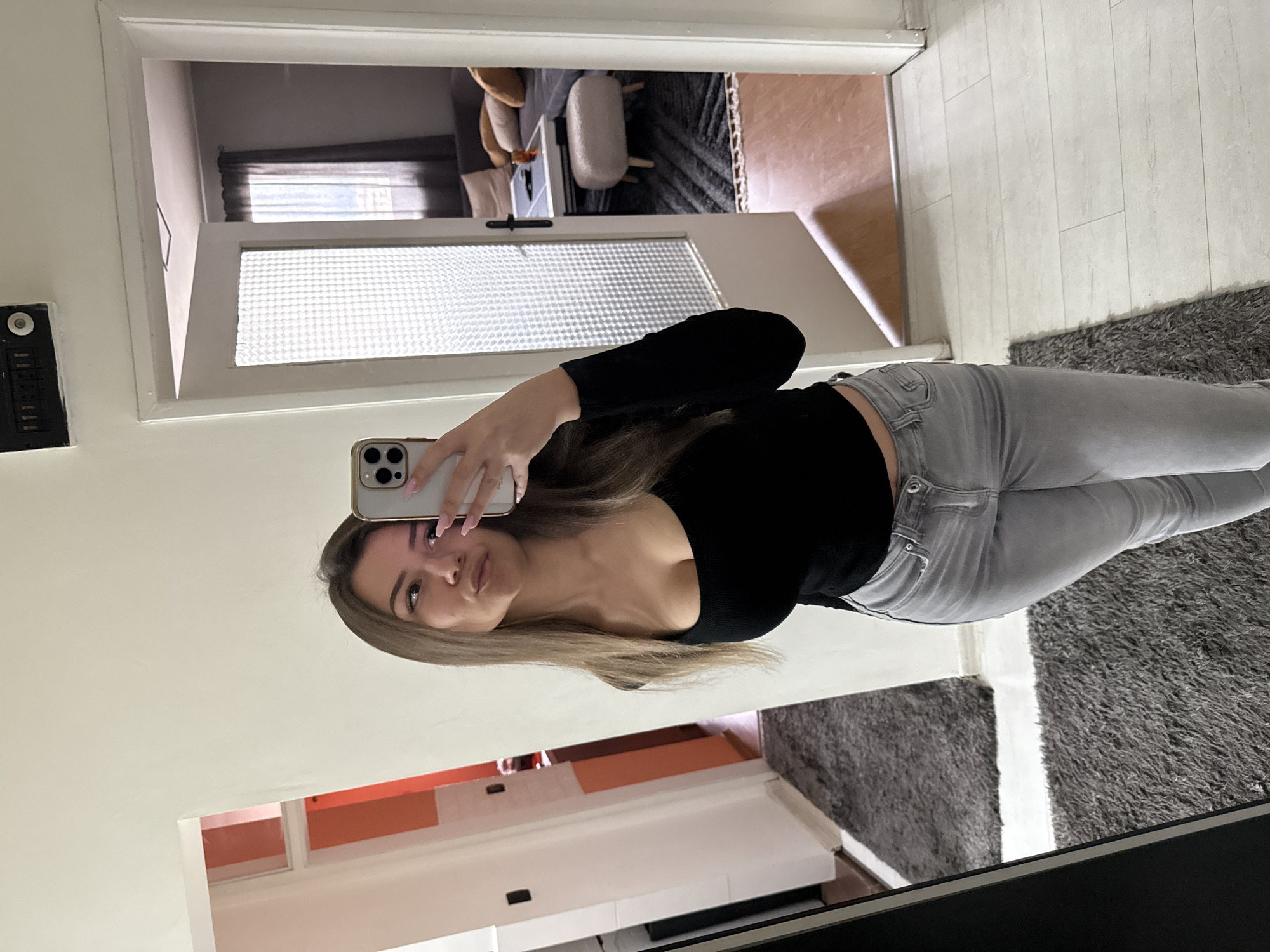 A young woman is taking a selfie in a mirror. She has long blonde hair and is wearing a black top and grey jeans. The background appears to be a modern apartment. The mirror reflects a hallway with a patterned rug and a view into another room with a chair and curtains. A white cabinet is visible in the lower portion of the reflection. The location cannot be determined from the image.