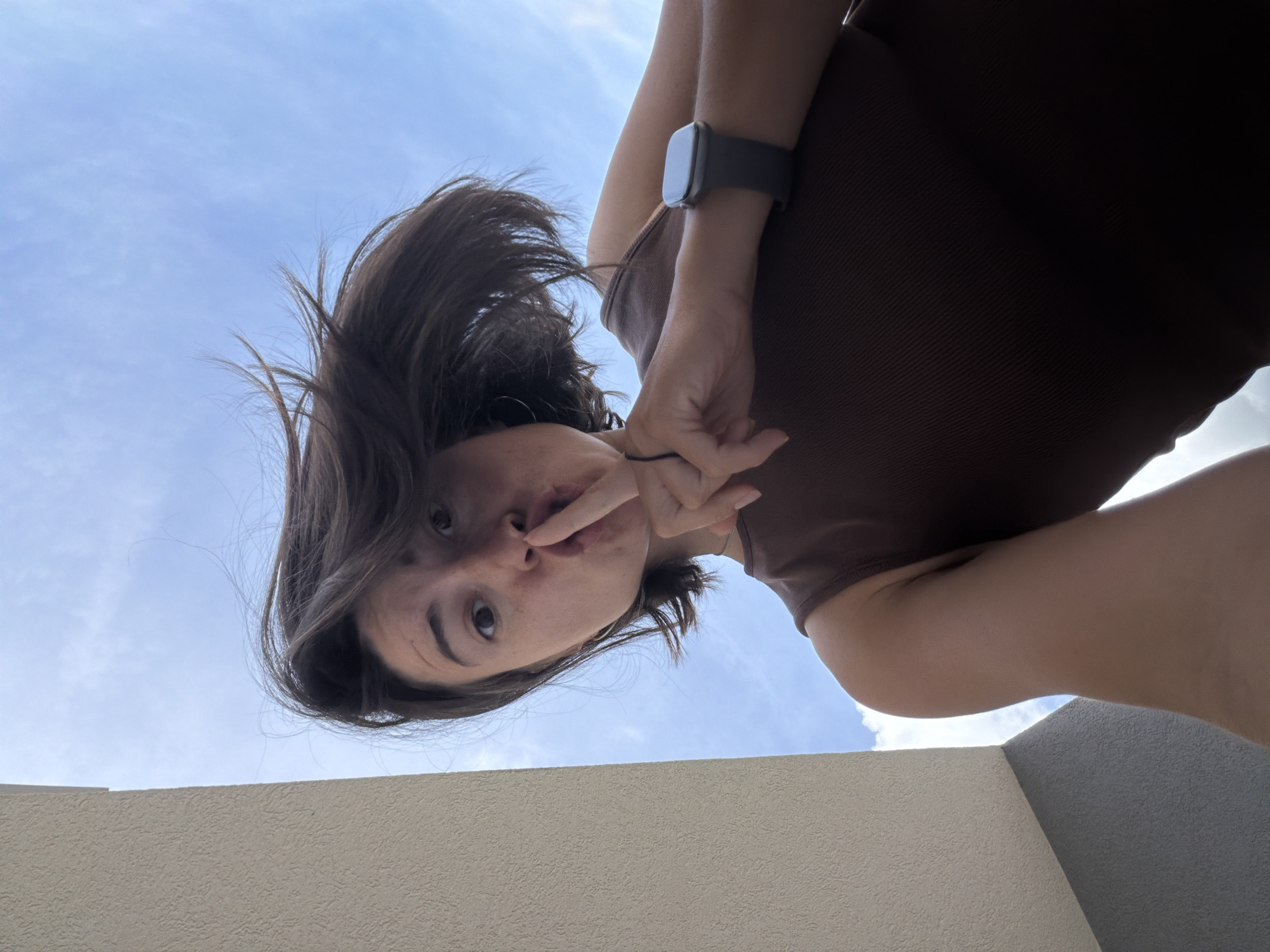 A woman with dark brown hair stands on a concrete balcony against a backdrop of a partly cloudy, light blue sky. She wears a dark brown ribbed top and an Apple Watch on her left wrist. Her left hand is raised, with her index finger over her lips in a shushing gesture. The camera angle is looking up at her from below, and the location appears to be a balcony in Budva, Montenegro, based on typical architectural styles and environmental conditions.
