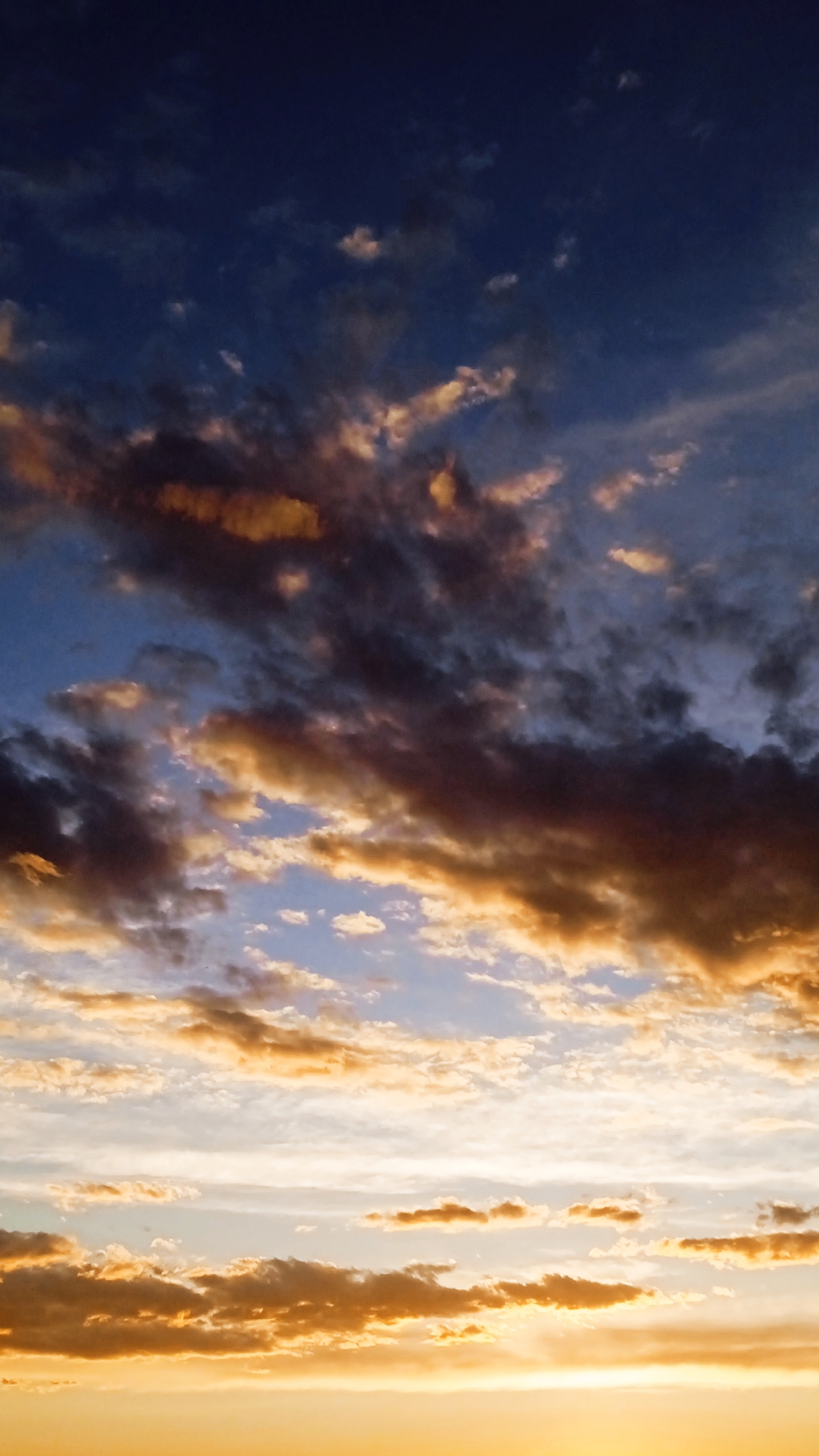 This is a vertical, medium shot of a dramatic sunset or sunrise sky. The scene is entirely composed of the sky, with no discernible ground or horizon line. The clouds are varied, with some appearing dark and heavy, tinged with deep blues and purples, while others are illuminated by the sun's rays, glowing with vibrant oranges, yellows, and pinks. The light from the setting or rising sun creates a gradient across the sky, transitioning from a deep, almost blackish blue at the top to a bright, warm yellow at the bottom. The clouds are scattered and appear in layers, some wispy and ethereal, others more dense and textured. The overall impression is one of a powerful and dynamic natural display, evoking a sense of awe and perhaps a touch of melancholy or peace. The time of day is clearly indicated by the warm, directional light and the colors of the sky. There is no visible text or any human or animal subjects.