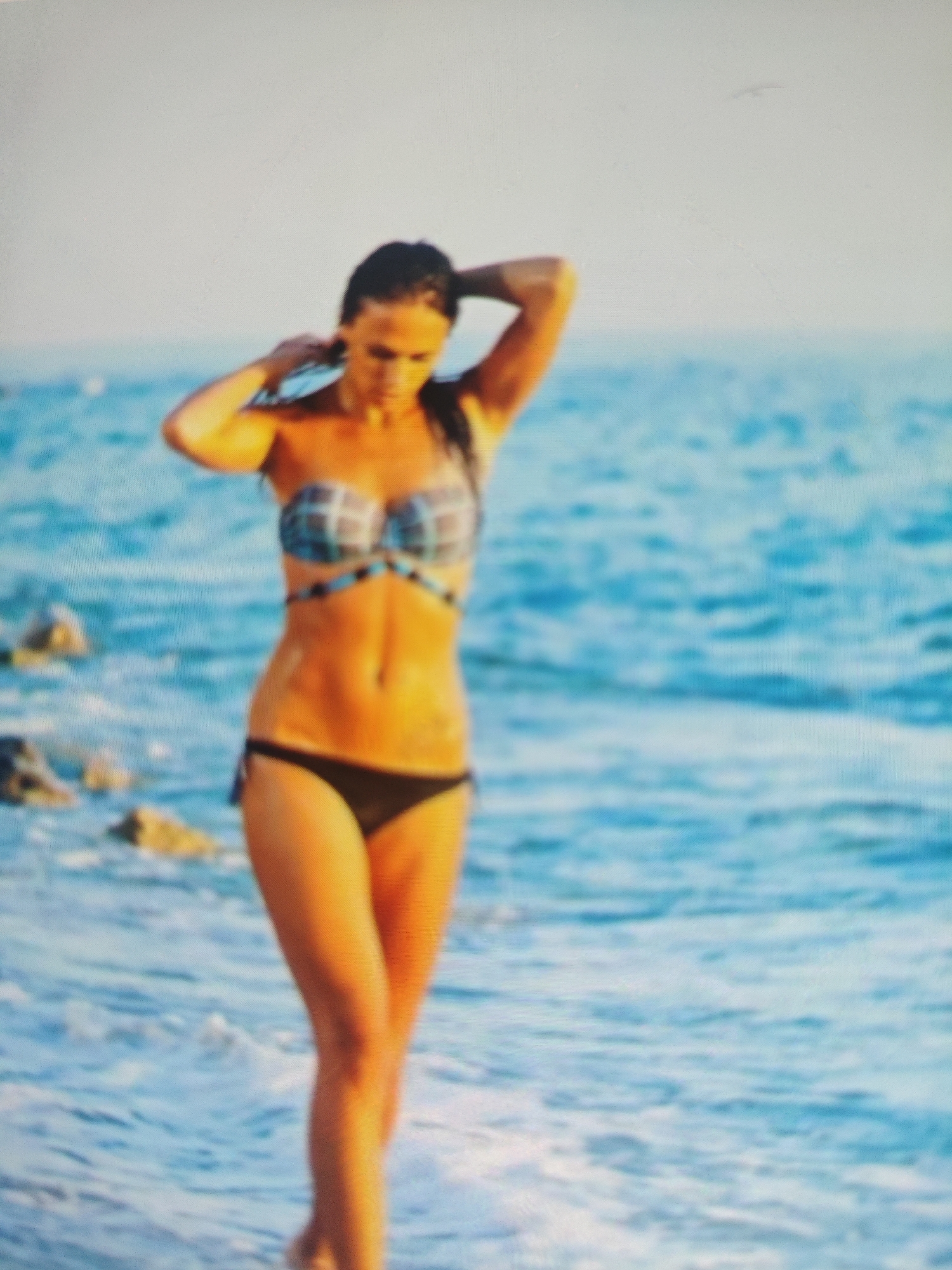 A woman in a bikini stands on a beach with the ocean behind her. The woman is facing to the left, with her arms raised to her head. The bikini top is checkered blue and white, and the bikini bottom is black. She has long, dark hair. The water is blue and choppy, with a few rocks visible in the foreground. The sky is a pale blue. This image does not depict any location in the city of Krasnaya Polyana, Russia.