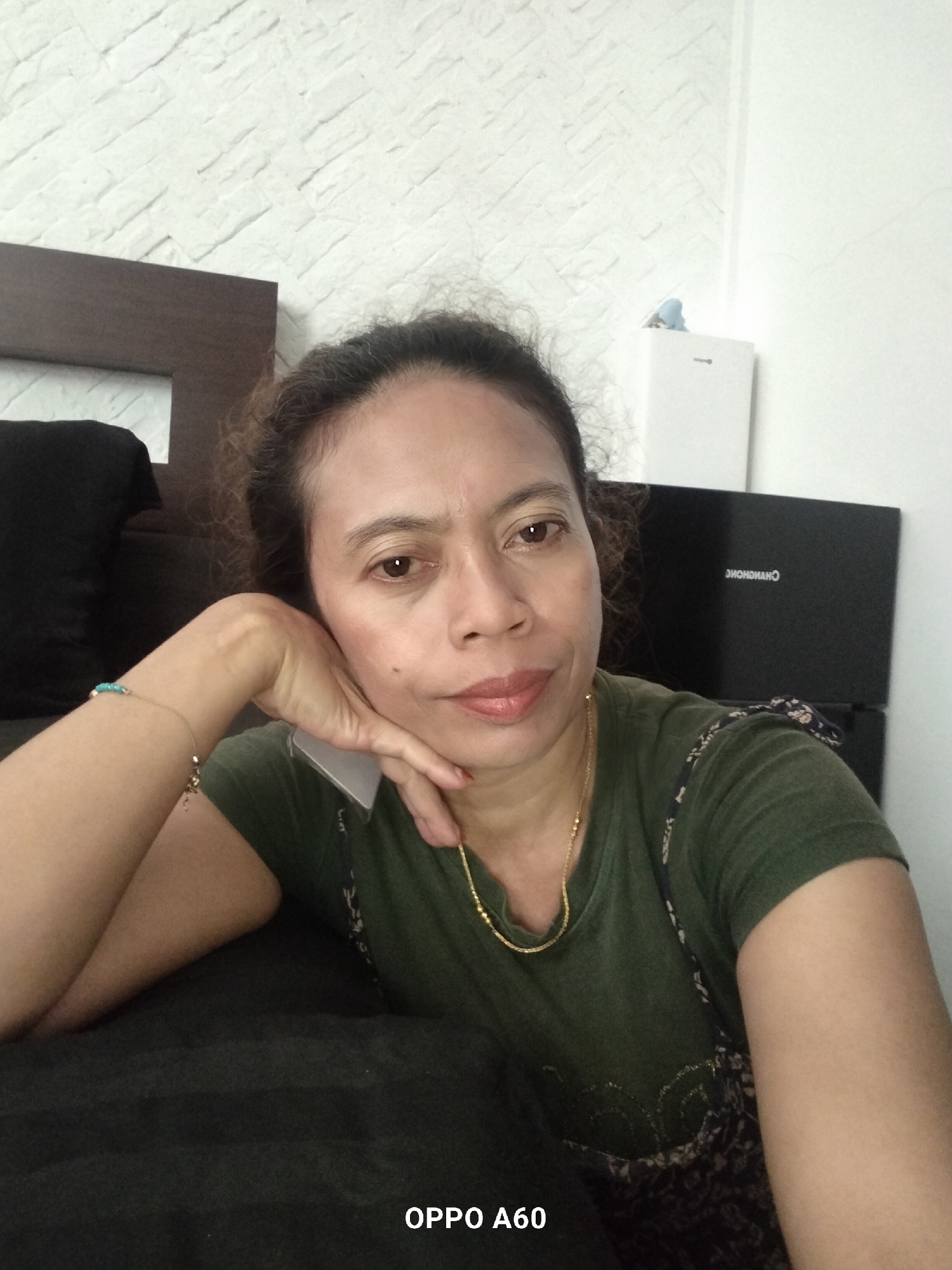 An indoor, eye-level, medium selfie shows a Filipino woman with brown curly hair, brown eyes, and fair skin posing for a photo. The woman is wearing a green t-shirt and a gold necklace. Her left arm is resting on a black couch, with her hand supporting her chin. A turquoise bracelet is on her left wrist. The background consists of a white brick-patterned wall, a dark brown headboard, and a black box with white text. The text reads "CHONOHONCH." The bottom of the image shows "OPPO A60." The location is in North Kuta, Indonesia.