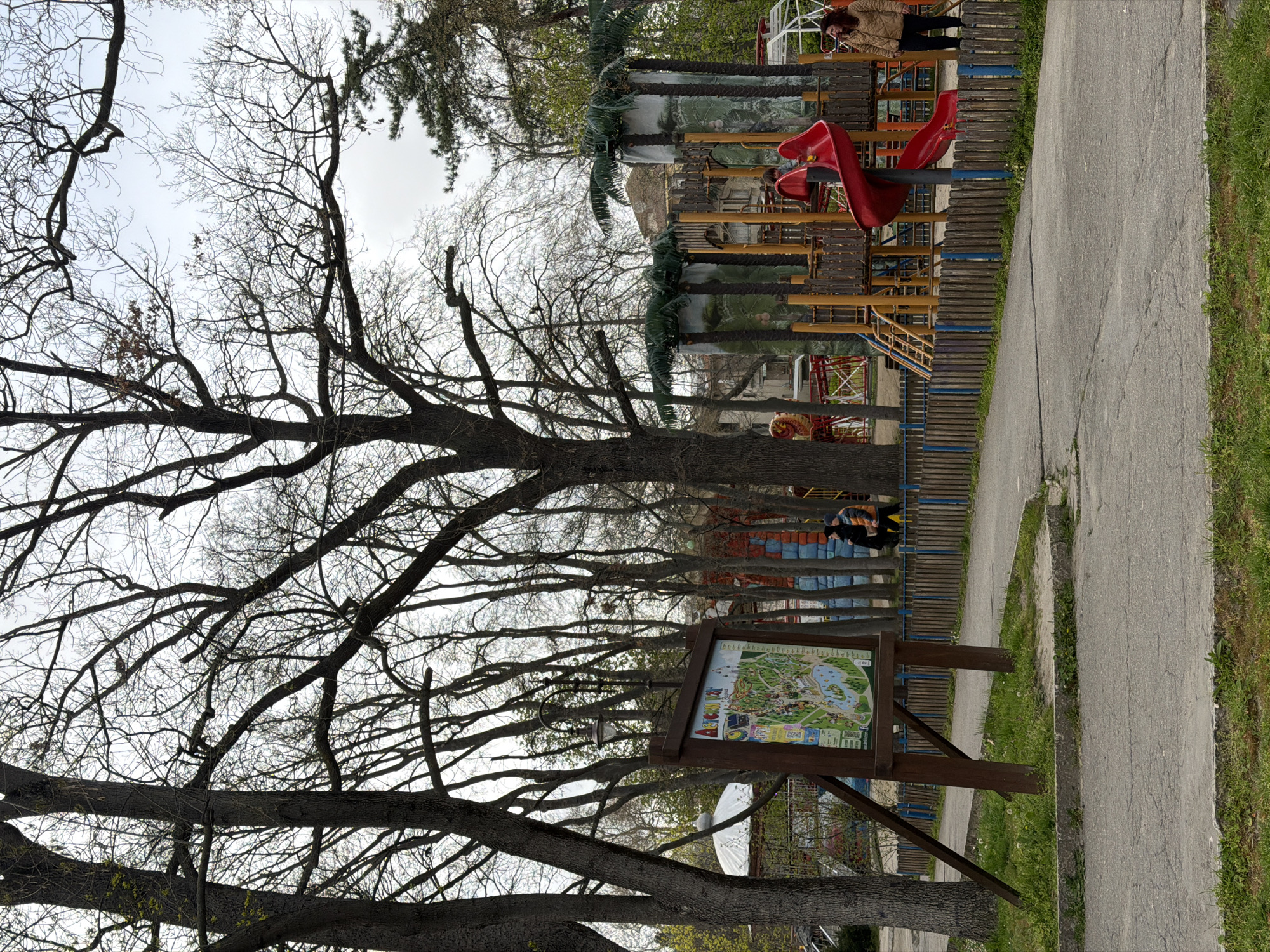 The image shows a section of Primorski Park in Varna, Bulgaria. A paved path runs along the right edge of the frame. To the left of the path is a wooden fence, behind which is a playground area with a red slide and jungle-themed decorations. A park map is positioned near the path. Bare trees dominate the left side of the image, their branches filling the sky. A person is seen near the playground area. The sky appears overcast.