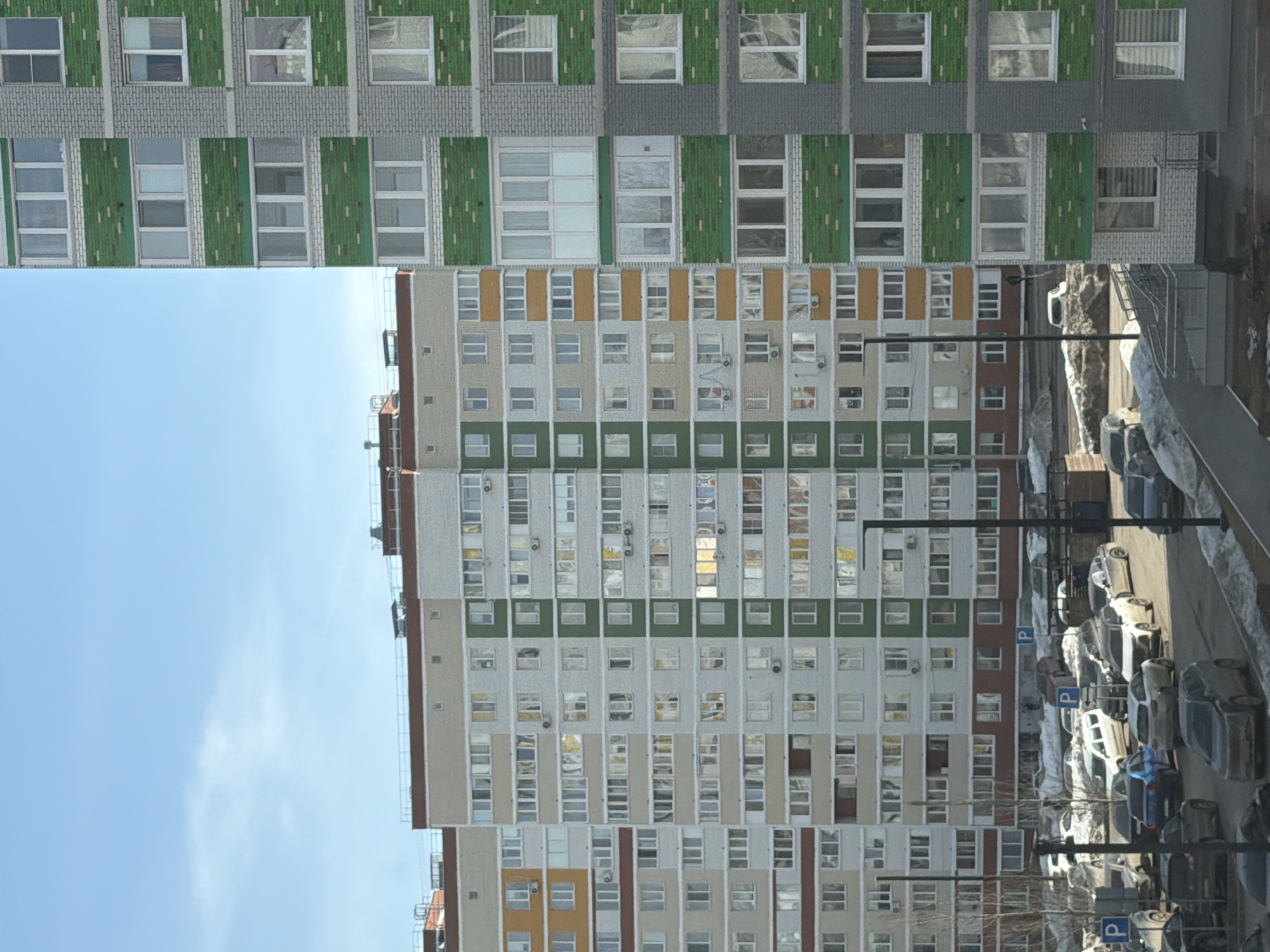 The image depicts an urban landscape in Izhevsk, Russia, featuring a high-rise residential building with a repetitive pattern of windows and colorful accents. The building is situated against a backdrop of a clear blue sky with scattered clouds. A street with parked cars runs parallel to the building, indicating a residential area. The architecture appears to be of a Soviet-era style, common in many Russian cities. There are no people visible in the scene.