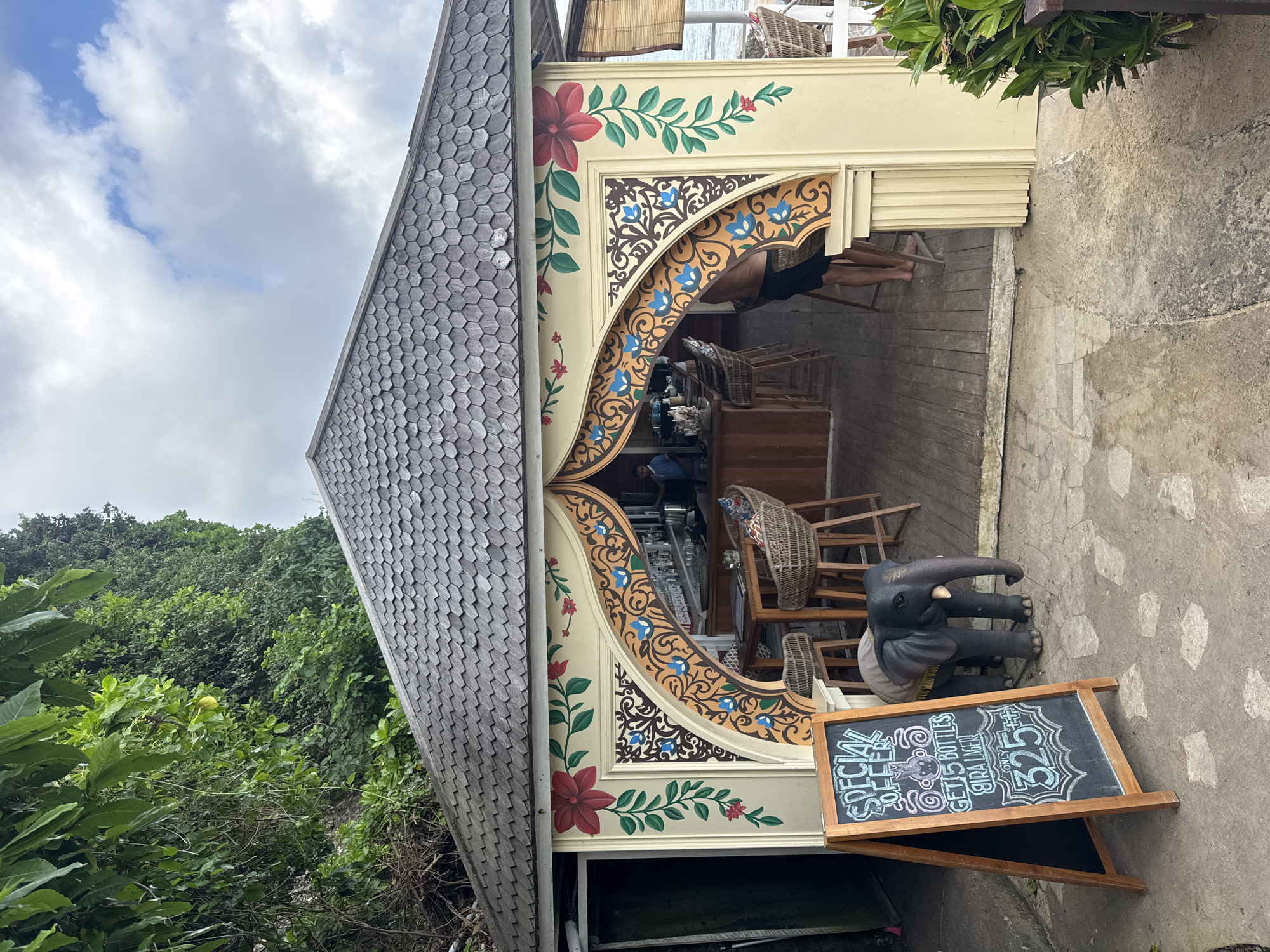 The image depicts an outdoor bar in Kuta Selatan, Indonesia. The bar's architecture features intricate floral and arabesque designs. Inside the bar, there are high wooden chairs and a person is visible. To the right of the bar's opening, there is a grey elephant statue and a blackboard sign displaying a special offer. The backdrop includes lush green foliage and a cloudy sky.