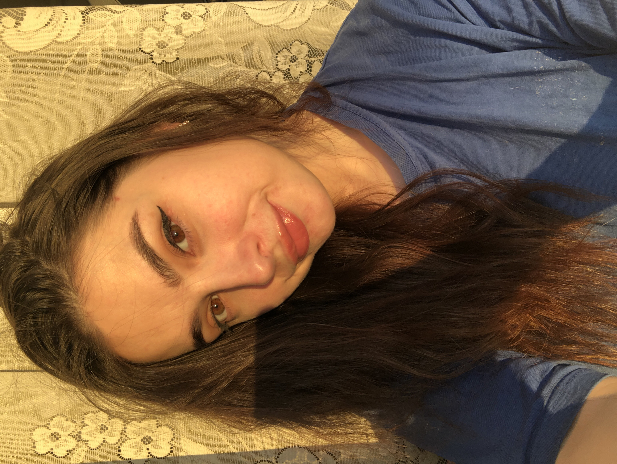 A young woman with long brown hair, brown eyes, and light skin is reclining on a surface covered with a white floral lace tablecloth. She is wearing a blue t-shirt and eyeliner. Sunlight illuminates the scene, casting shadows on her face and clothing. There are no indications about the location in the city of Noviy, Russia.