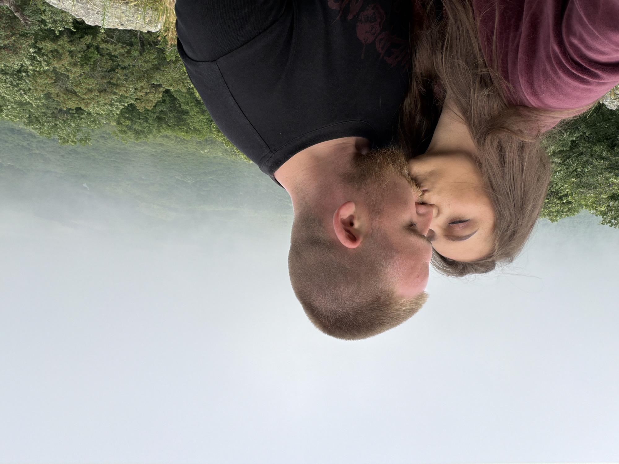 An intimate, close-up shot captures a man and a woman in a tender embrace, appearing to kiss or in very close proximity. The man, positioned on the left, has short, light brown hair and a beard, and is wearing a black t-shirt. The woman, on the right, has long, flowing brown hair and is dressed in a maroon long-sleeved shirt. Their heads are angled towards each other, with their eyes closed or mostly closed, conveying a sense of intimacy and affection.
The setting is an expansive outdoor landscape, likely a mountain or cliffside vista. Behind and slightly above the couple, a rocky ledge with sparse vegetation is visible, indicating they are positioned at an overlook. Below them, a vast, dense forest covers rolling hills or mountains, receding into the distance. The background is characterized by a soft, light mist or fog, particularly over the distant landscape, which blends into a pale, hazy sky. The natural lighting suggests it is daytime, possibly under an overcast sky, contributing to the soft, romantic atmosphere. No text is visible in the image.