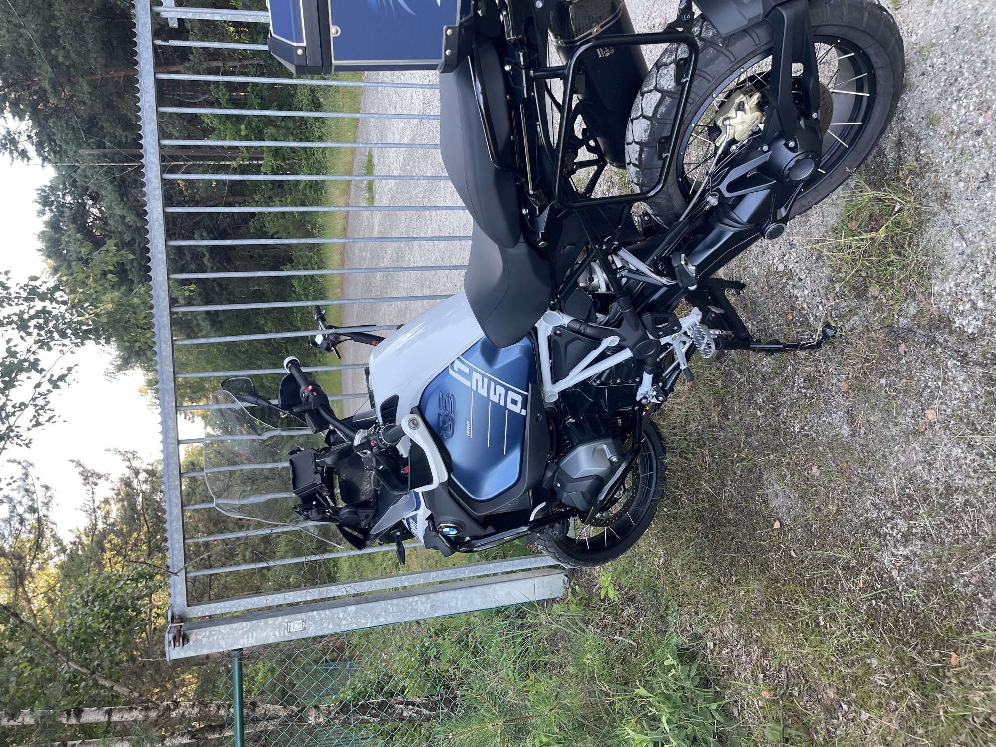 An outdoor, eye-level, medium shot shows a white and blue BMW R 1250 GS motorcycle parked on a gravel and grass area. The motorcycle is angled towards the right, with its kickstand down. The front of the motorcycle is to the left, featuring a clear windshield, handlebars with mirrors, and a black and silver front fender. The fuel tank is white and blue with "GS" branding. The rear of the motorcycle shows a black seat, black saddlebags, and a black exhaust system. Behind the motorcycle is a tall, grey metal gate with vertical bars, partially open. Beyond the gate is a dense forest with various trees. A green chain-link fence runs along the left side of the scene, bordering the motorcycle and the gate. The lighting suggests it is daytime, possibly late afternoon, with sunlight filtering through the trees.