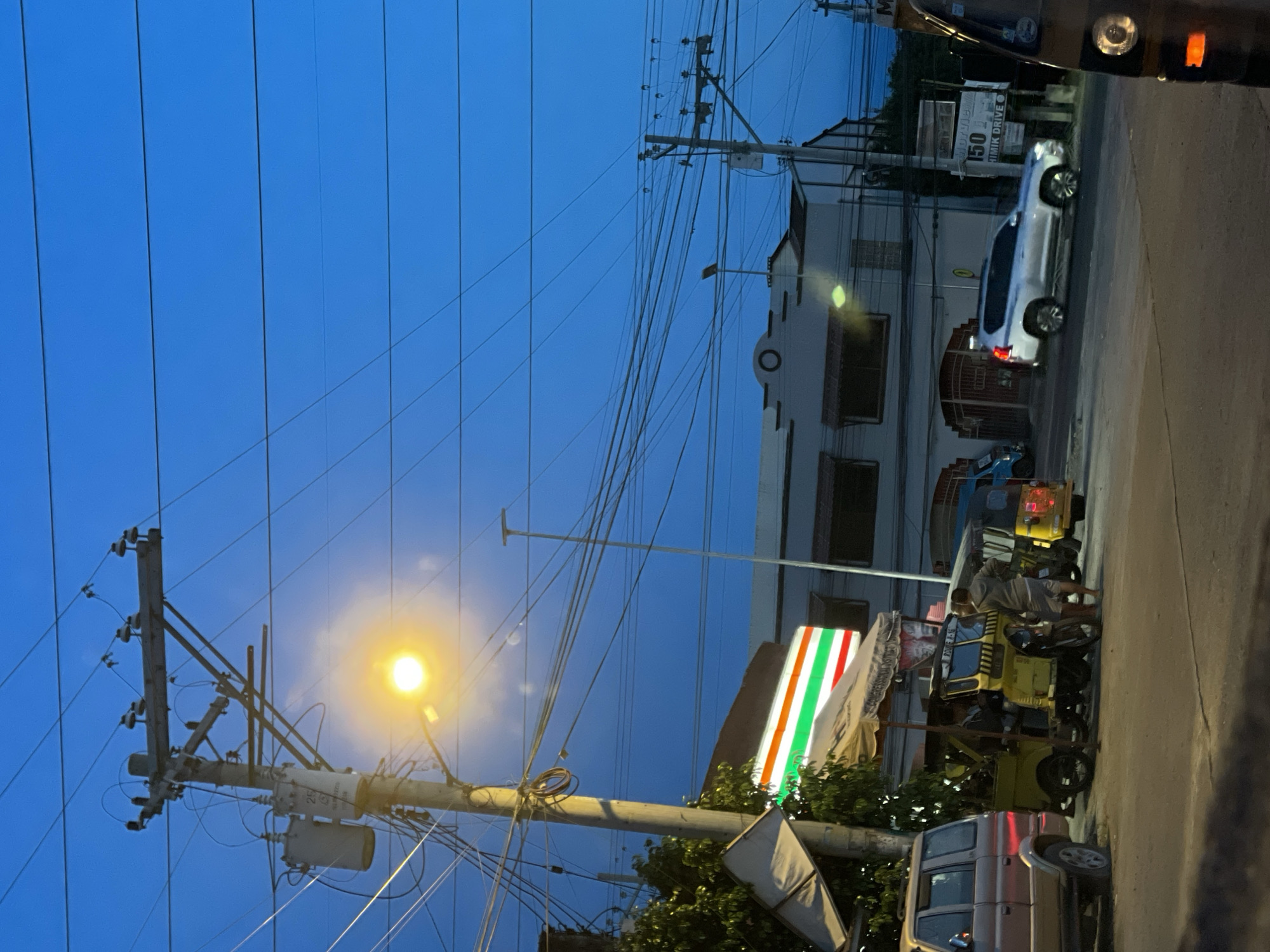 The image depicts a street scene in Davao City, Philippines, at dusk or early evening. The sky is a deep blue, indicating the transition from day to night. A prominent utility pole with numerous wires and a transformer stands to the left of the frame, with a bright streetlamp illuminating the immediate area.
In the background, there's a white building with visible windows and architectural details. A silver SUV is parked on the right side of the street. In front of a building, a colorful sign with red, green, and white stripes, likely a 7-Eleven logo, is visible.
Several vehicles, including what appears to be a yellow tricycle or three-wheeled motorcycle with a passenger, are parked or in motion on the street. A person is seen standing next to the yellow vehicle. A light brown pickup truck is parked in the foreground.
The scene suggests a typical urban environment in the Philippines with the presence of common modes of transportation and commercial establishments. The time of day is marked by the twilight sky and the illuminated streetlamp. No specific event or emotional cues are evident, making it a snapshot of everyday life. Some faint text is visible on a sign behind the silver SUV, reading "150 DRIVEMIK DRIVE".