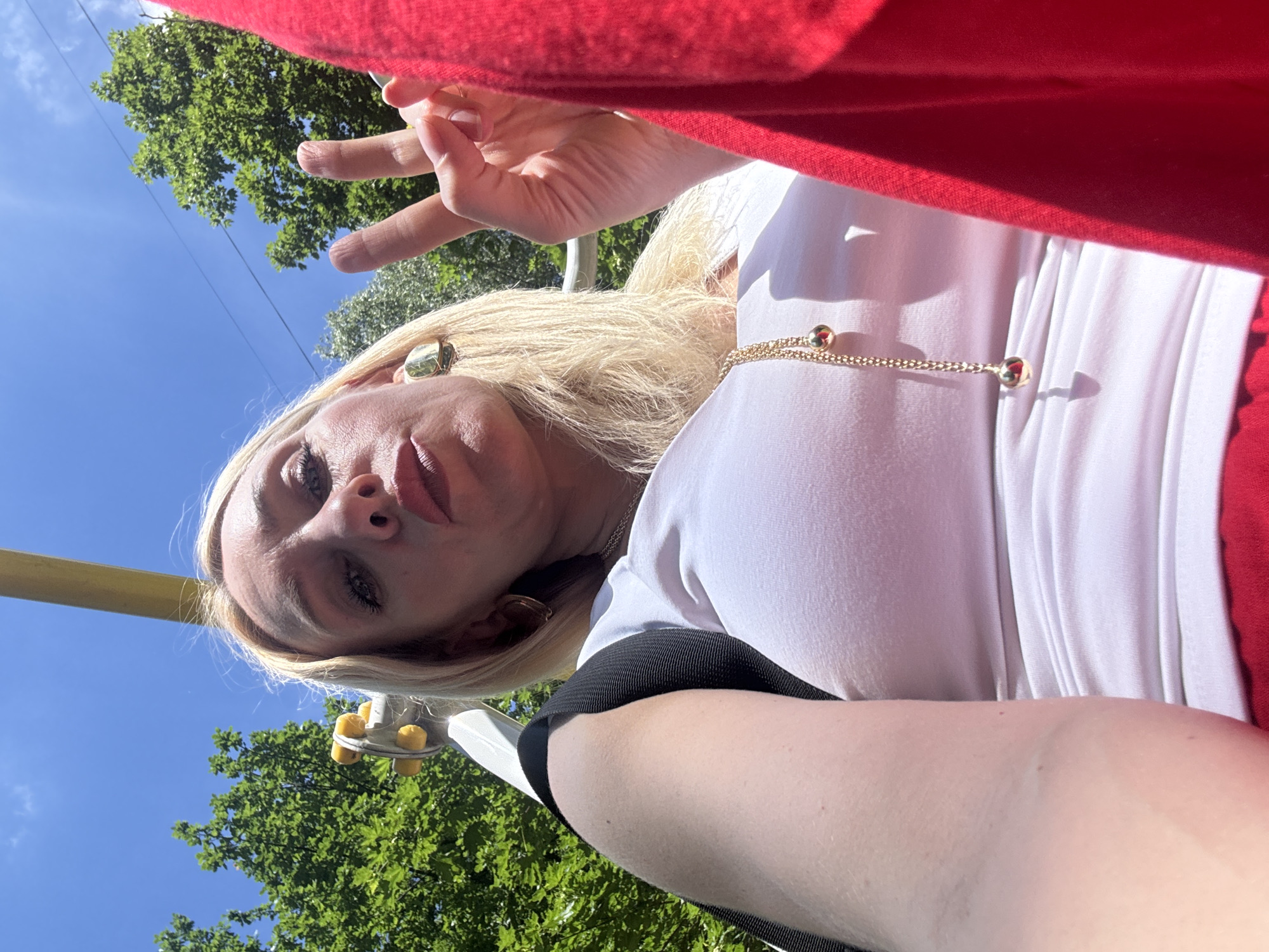 In this close-up, low-angle selfie taken outdoors on a sunny day, a blonde woman is prominently featured against a backdrop of green trees and a bright blue sky. She is looking directly at the camera with her mouth slightly open. She is wearing a white t-shirt and red bottoms, with a gold necklace adorned with two small spheres visible. Her right hand is raised, displaying a peace sign. She is also wearing large, square-shaped gold earrings. Part of a black strap and a yellow bar are visible in the upper left corner, suggesting she may be on a swing set or similar amusement park equipment. The overall impression is a candid, upbeat moment captured in an outdoor setting.