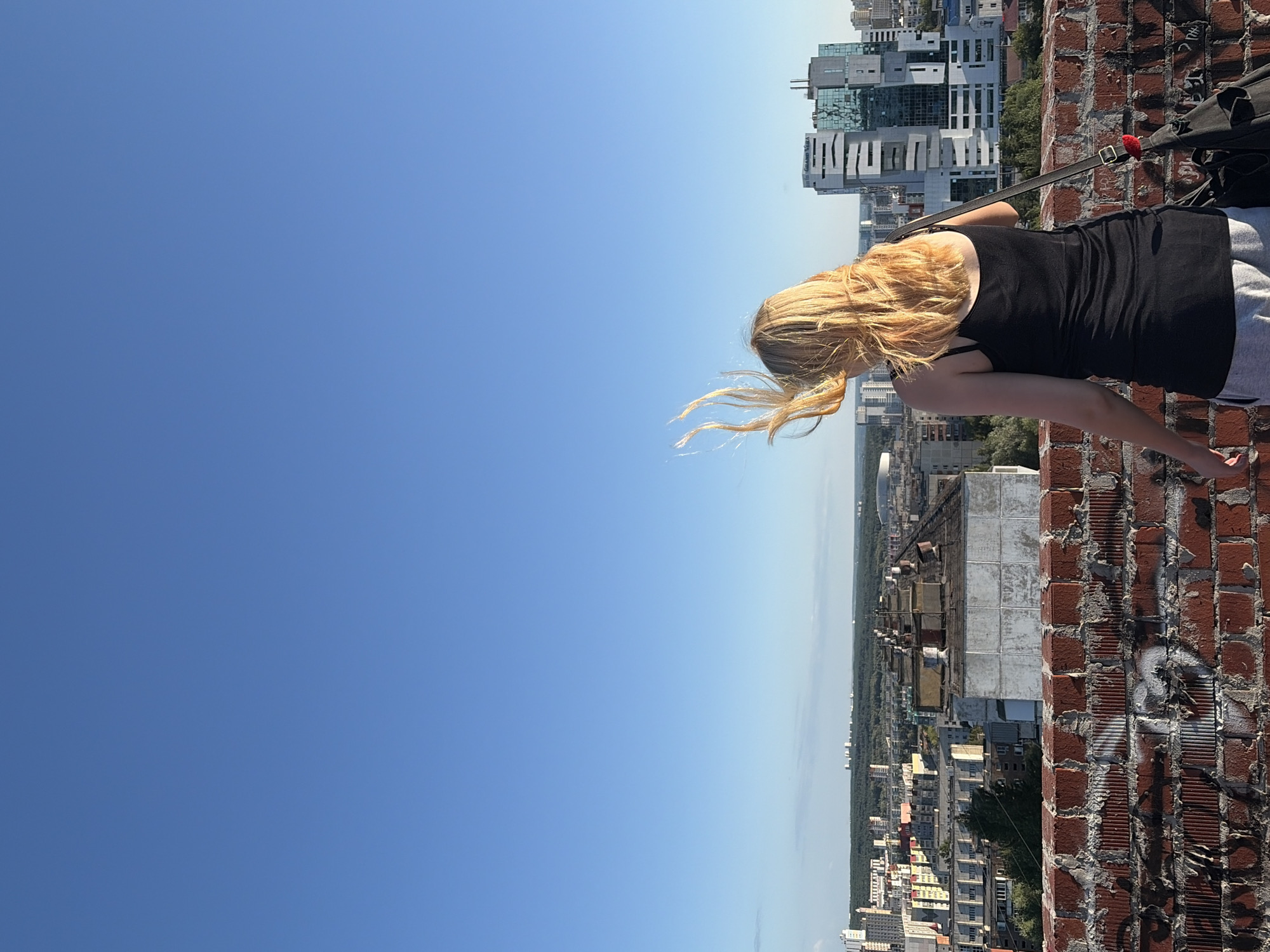 A young woman with blonde hair, viewed from behind, stands on a rooftop overlooking a city. Her hair is blowing in the wind, and she is wearing a black tank top and gray sweatpants. She has a black bag slung over her shoulder. In the background, a cityscape unfolds with various buildings, including modern high-rises. The sky is a clear, vibrant blue. This vantage point is consistent with the rooftop of a building in the area of Chelyabinsk, Russia, known for its mix of older and newer urban architecture. The foreground features a brick wall with graffiti.