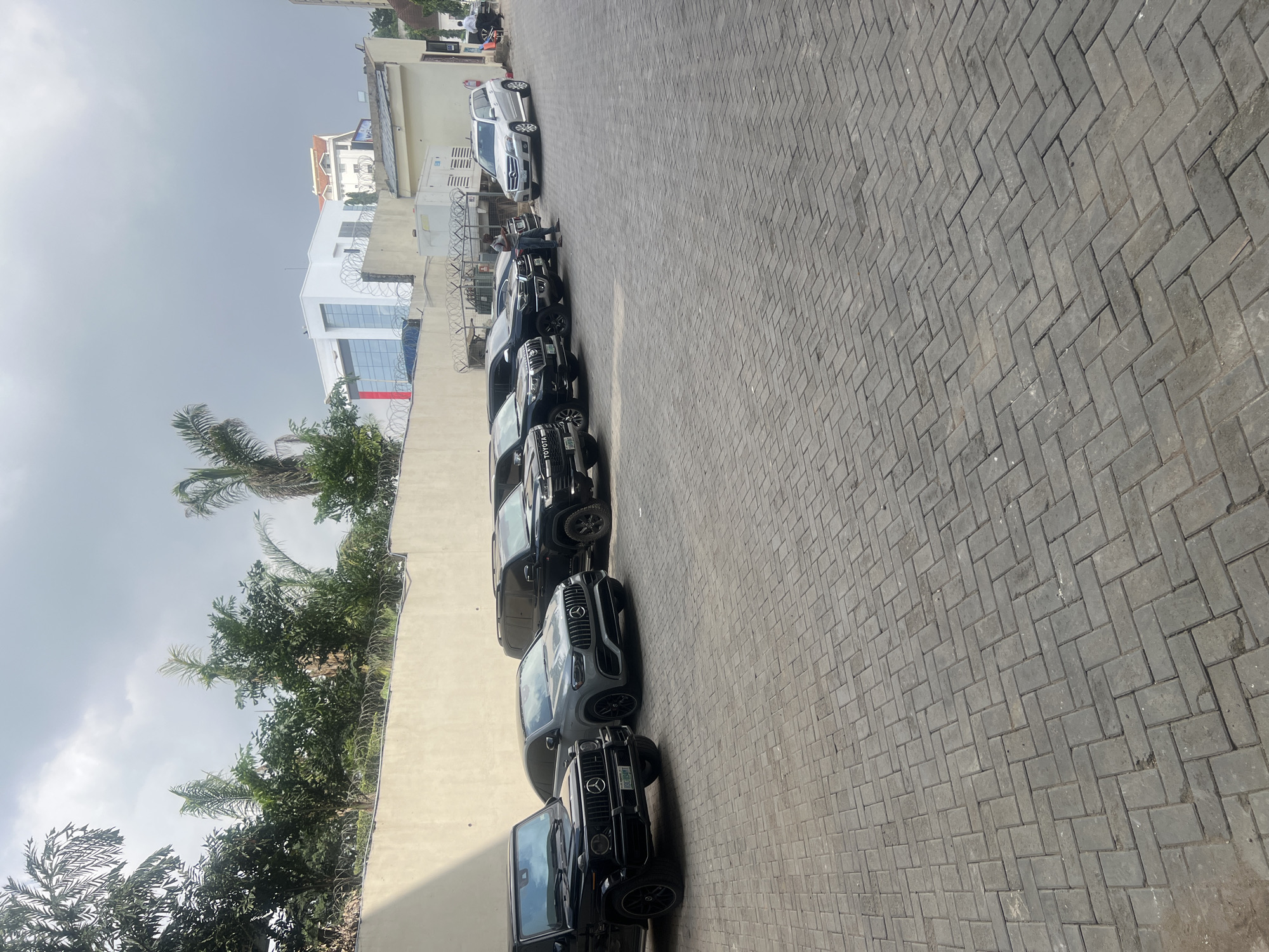 In the city of Abuja, Nigeria, a paved lot adjacent to a light-colored wall features a line of parked vehicles. From left to right, a black Mercedes-Benz G-Wagon is parked closest to the wall. To its right is a dark grey Mercedes-Benz GLE Coupe. Further down the line are several black SUVs, including a Toyota Land Cruiser. A silver Mercedes-Benz GLK-Class is parked further to the right, with a man standing near it, looking at his phone. Another man is visible further down in the background, standing near a white generator unit. Behind the vehicles, the wall is topped with barbed wire, and beyond that, lush green trees and buildings are visible under a cloudy sky. The ground is paved with grey interlocking bricks.