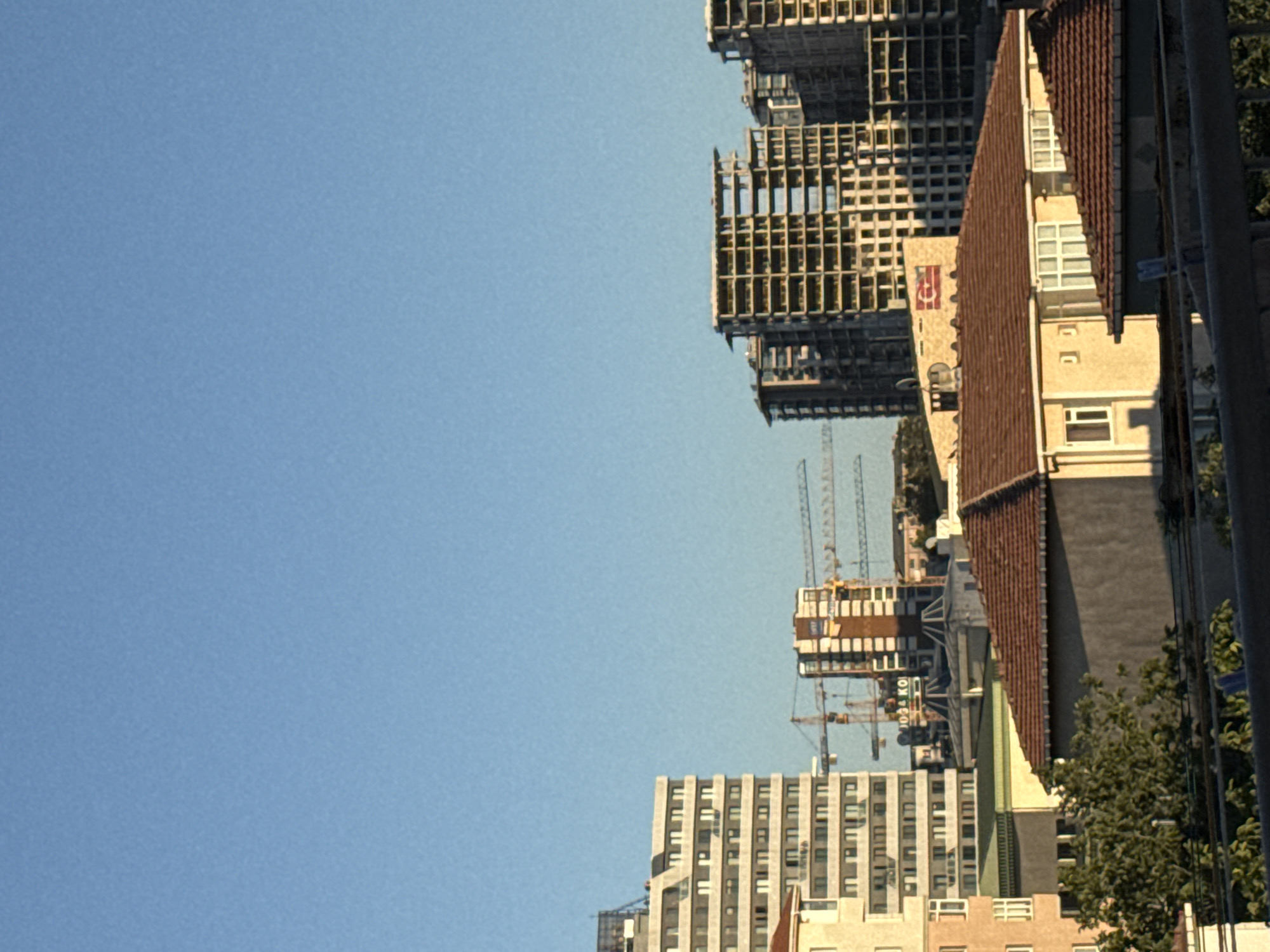 The image captures a cityscape with several buildings and construction cranes under a clear blue sky, likely taken in Gungoren, Turkey. In the foreground, there are residential-style buildings with terracotta tiled roofs. Behind them, taller apartment buildings are visible, some appearing to be completed and others under construction. The unfinished buildings reveal their concrete skeletal structures. Several construction cranes, in yellow and red, are positioned among the buildings, indicating ongoing development. The overall scene suggests a dynamic urban environment in a state of growth.