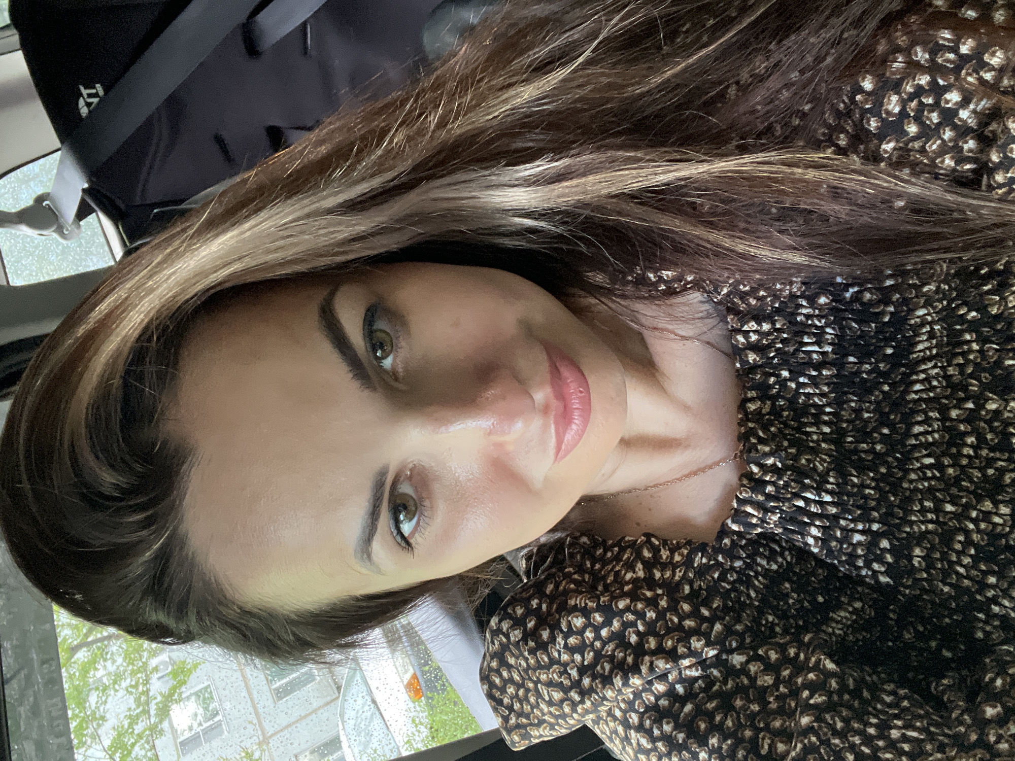 A woman is taking a selfie in a car. She has long, wavy brown hair with blonde highlights and is wearing a dark brown and tan animal-print top. The background shows a window with raindrops on it, a building, and trees. A car seat with a black seatbelt is visible behind her.