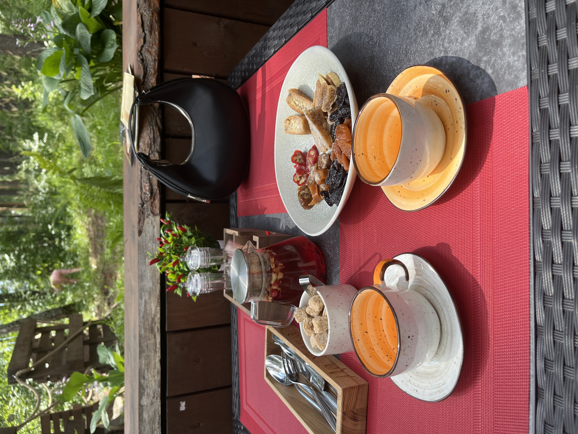 This image depicts an outdoor dining setting, likely a cafe or restaurant, with a natural, wooded background. The table is set with two orange and white speckled ceramic cups and saucers, a glass teapot filled with red liquid and fruit pieces, a small white dish of brown sugar cubes with a silver scoop, and a wooden cutlery holder containing silver utensils. A plate of dried fruits, nuts, and what appear to be dried chili peppers is also visible. To the rear of the table, a black handbag rests on a wooden surface. The environment suggests a leisurely meal or break, with greenery and trees in the background, implying a location outside of a dense urban area. A blurred figure of a person is visible in the background, walking through the trees. While Chebarkul is a city in Russia, the specific details in the image do not provide definitive evidence to confirm or deny this location.