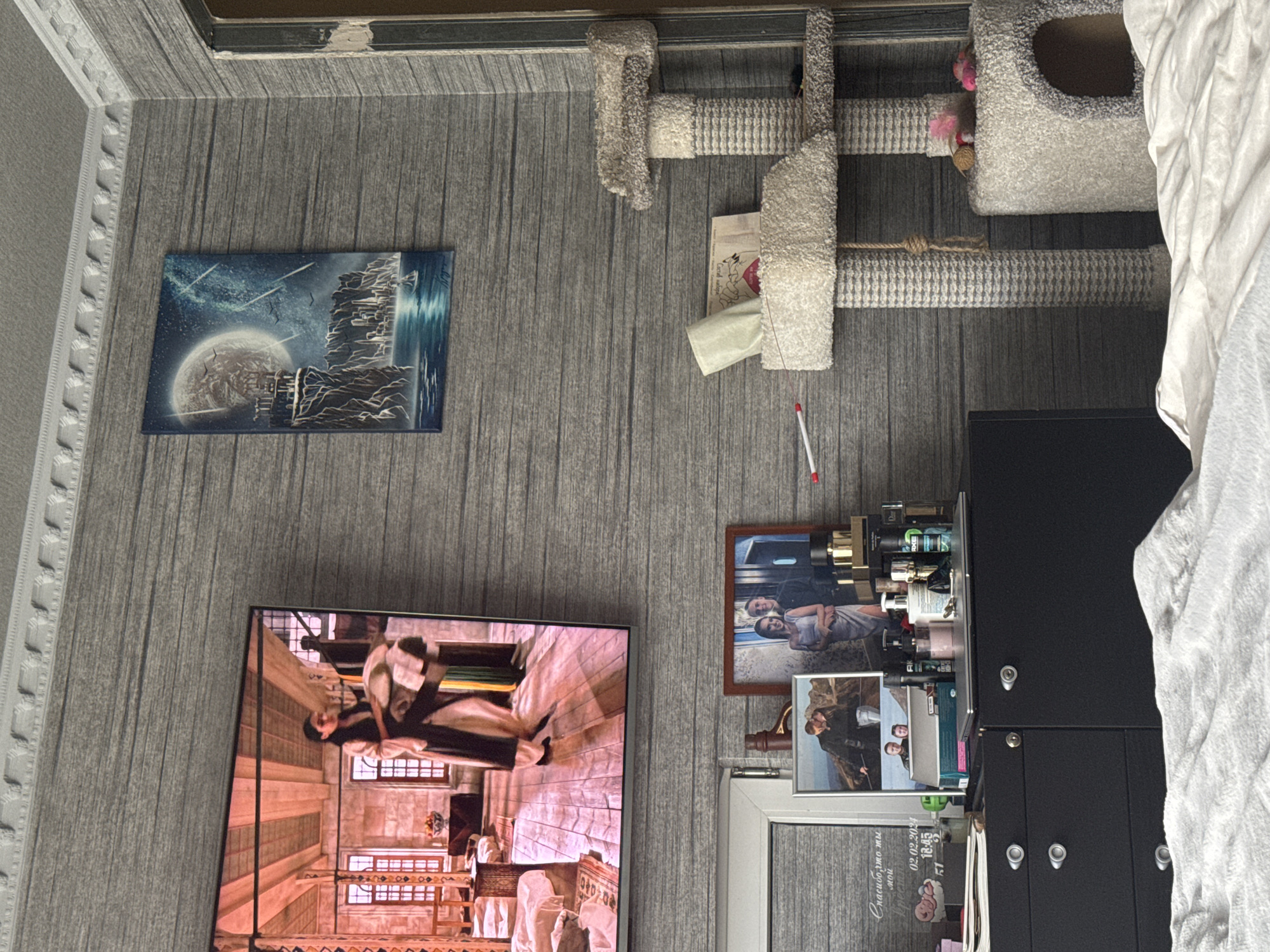 This is a medium shot of a domestic interior, likely a bedroom, in Makiivka, Ukraine. The scene is dominated by a wall with horizontal grey textured wallpaper. On the left, a large flat-screen television displays a scene of individuals in traditional clothing within an ornate interior. Above the television, decorative molding is visible on the ceiling.
To the right of the television, a framed artwork depicting a fantastical landscape with a moon, a city, and water is mounted on the wall. Further to the right, a multi-level cat tree made of sisal rope and plush fabric stands against the wall, with a small cat bed at its base.
Below the television, a black cabinet with silver drawer pulls is present. On top of the cabinet, various items are arranged, including framed photographs of couples, perfume bottles, and a laptop. A white window frame is visible on the left side of the cabinet, with some text inscribed on it. The overall impression is of a lived-in space with personal decorative elements.