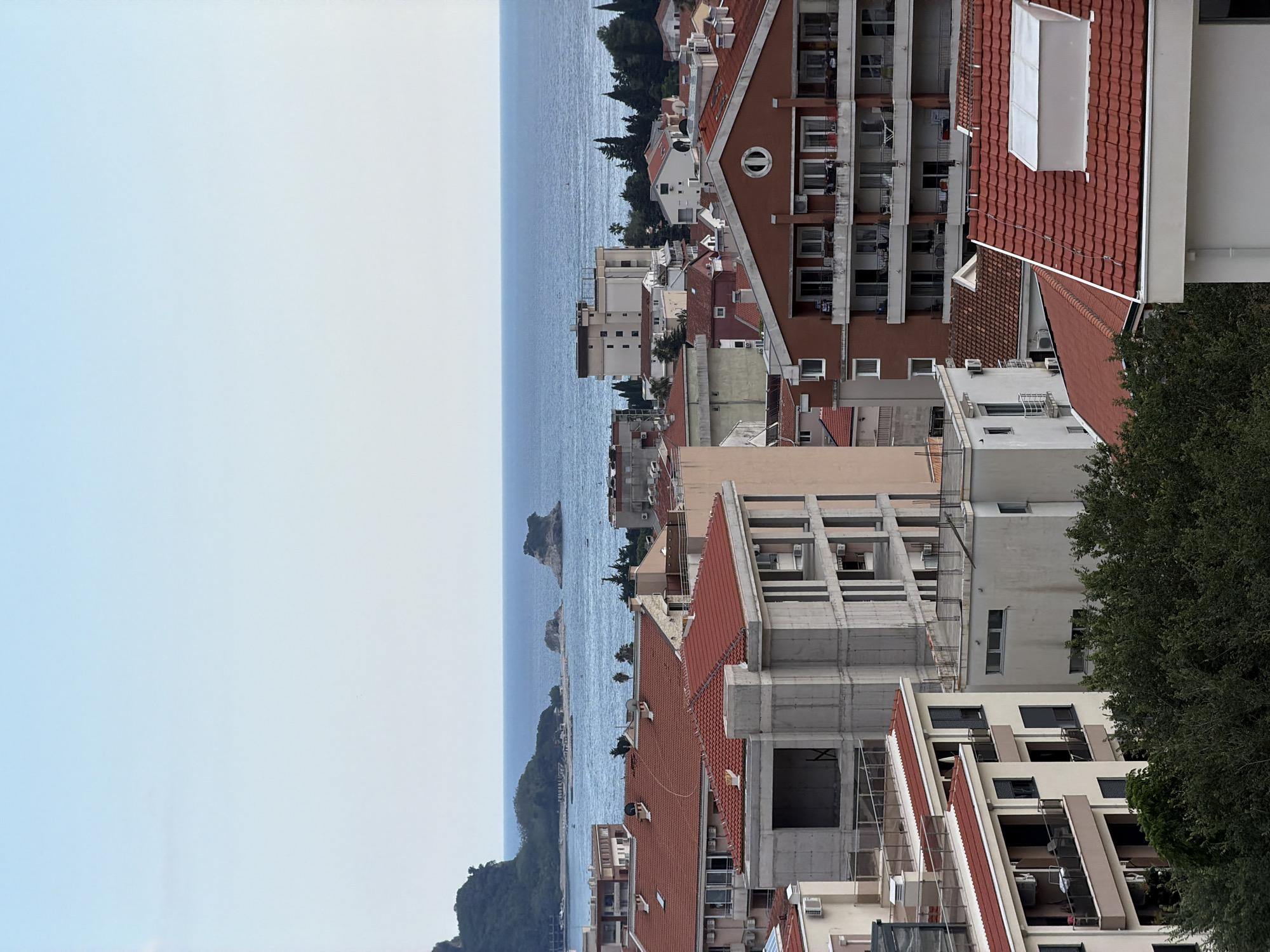 This is an outdoor, eye-level, vertical, medium shot of a coastal city, likely Budva, Montenegro, with the Adriatic Sea visible in the background. The foreground is dominated by a dense cluster of buildings with red tiled roofs and light-colored facades. Some buildings appear to be residential apartments with balconies, while others are under construction, showing exposed concrete structures and rebar.
In the mid-ground, the buildings continue, with some featuring light beige and white exteriors. One prominent building is a dark red brick structure with multiple floors of balconies. Further back, the coastline is lined with more buildings, some appearing older with terracotta roofs, and trees.
The background features the calm, blue Adriatic Sea, with several small rocky islands scattered on its surface. A distant, hazy green coastline can be seen on the left, and the sky is a pale, clear blue. The overall impression is of a bustling coastal town with ongoing development. No people are visible in the image.
