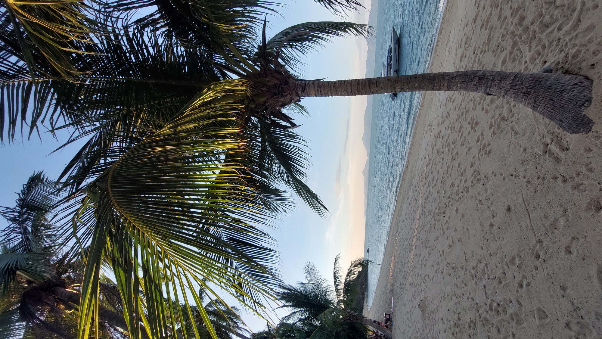 The image depicts a tropical beach scene in Baan Mae Nam, Thailand. The foreground is dominated by palm trees with green fronds, some partially obscuring the view of the sky. The beach stretches horizontally, with light-colored sand covered in numerous footprints. The ocean is visible beyond the beach, with calm, light blue water. A small boat is anchored near the shore. A distant mountain range is visible on the horizon under a clear sky. People are present in the lower right corner. The image is slightly tilted.