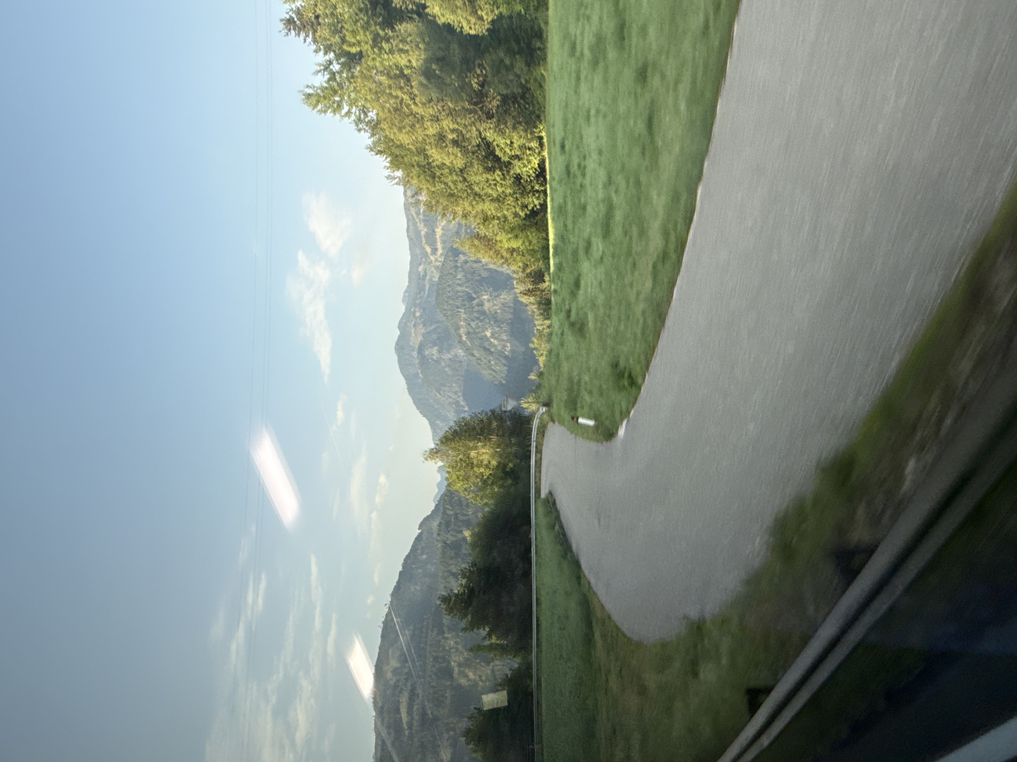 This is a photograph of a winding road in a mountainous landscape. The road is paved and curves to the right in the foreground. On either side of the road are grassy slopes. In the background, there are forested mountains under a clear blue sky with some scattered clouds. There are reflections visible on the surface of the image, suggesting it was taken through a window. There are no people or animals visible in the image.